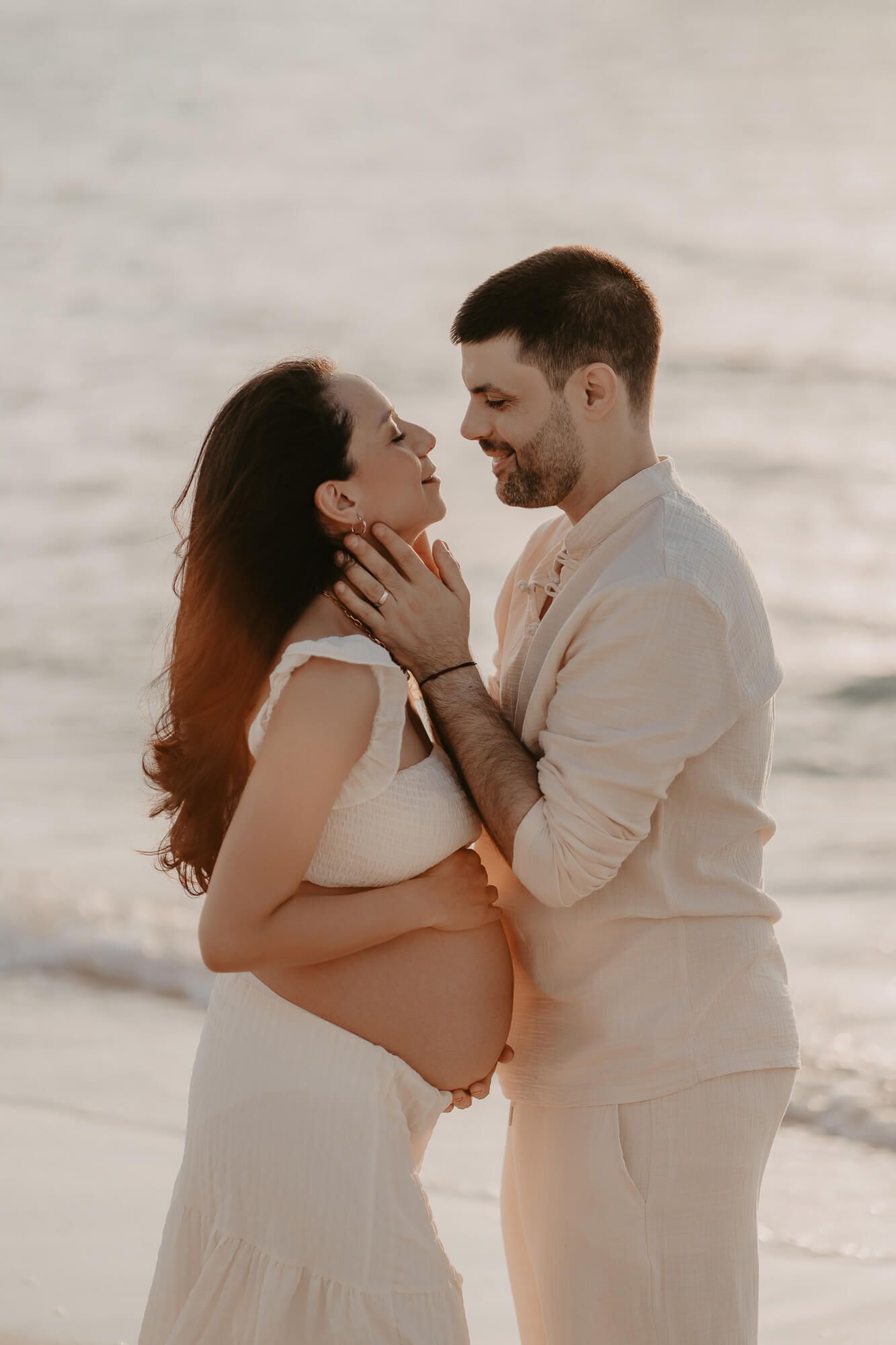Romantic maternity photo of a couple in white outfits embracing on the beach at sunset.