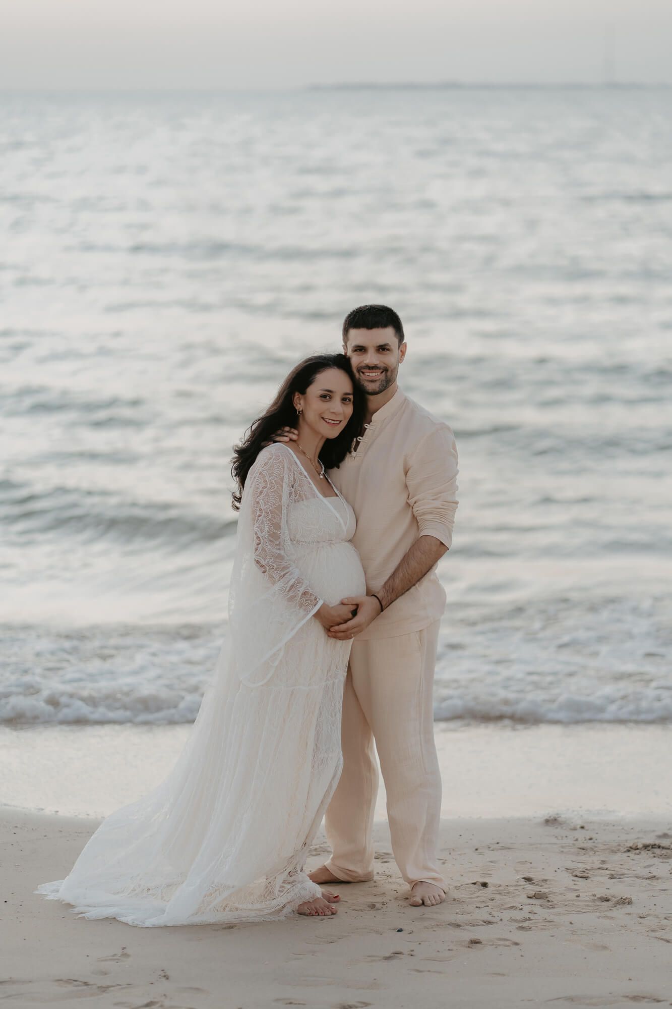 A couple stands together on a beach at sunset wearing light-colored formal attire.