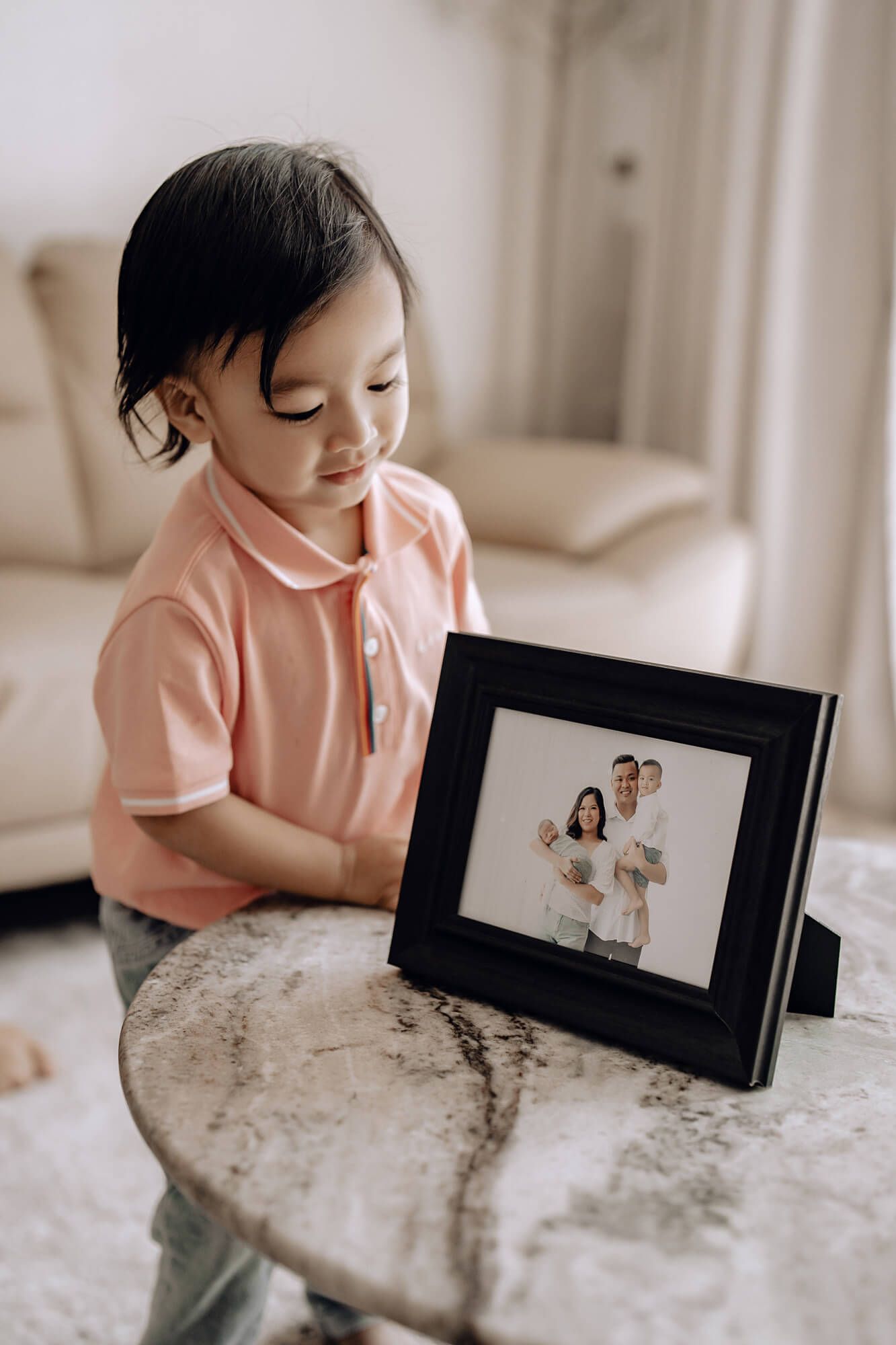 A small child leans over a table to look at a black picture frame displaying family photos in a bright room.
