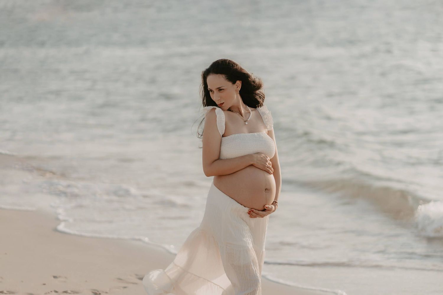 A pregnant woman in a white dress walks gracefully along a beach shoreline during a serene sunset photoshoot.
