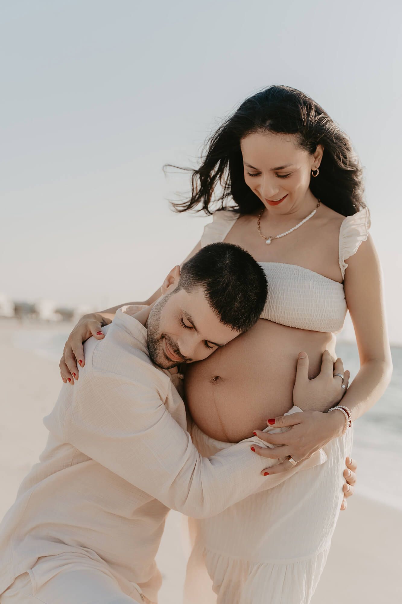 Intimate maternity photo on beach shows couple in white clothing embracing growing baby bump.