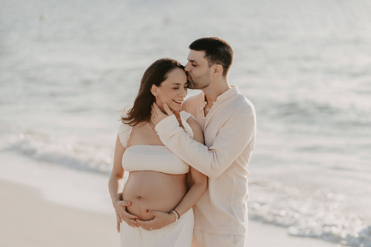 A tender maternity photoshoot on the beach with couple embracing by the ocean at sunset.