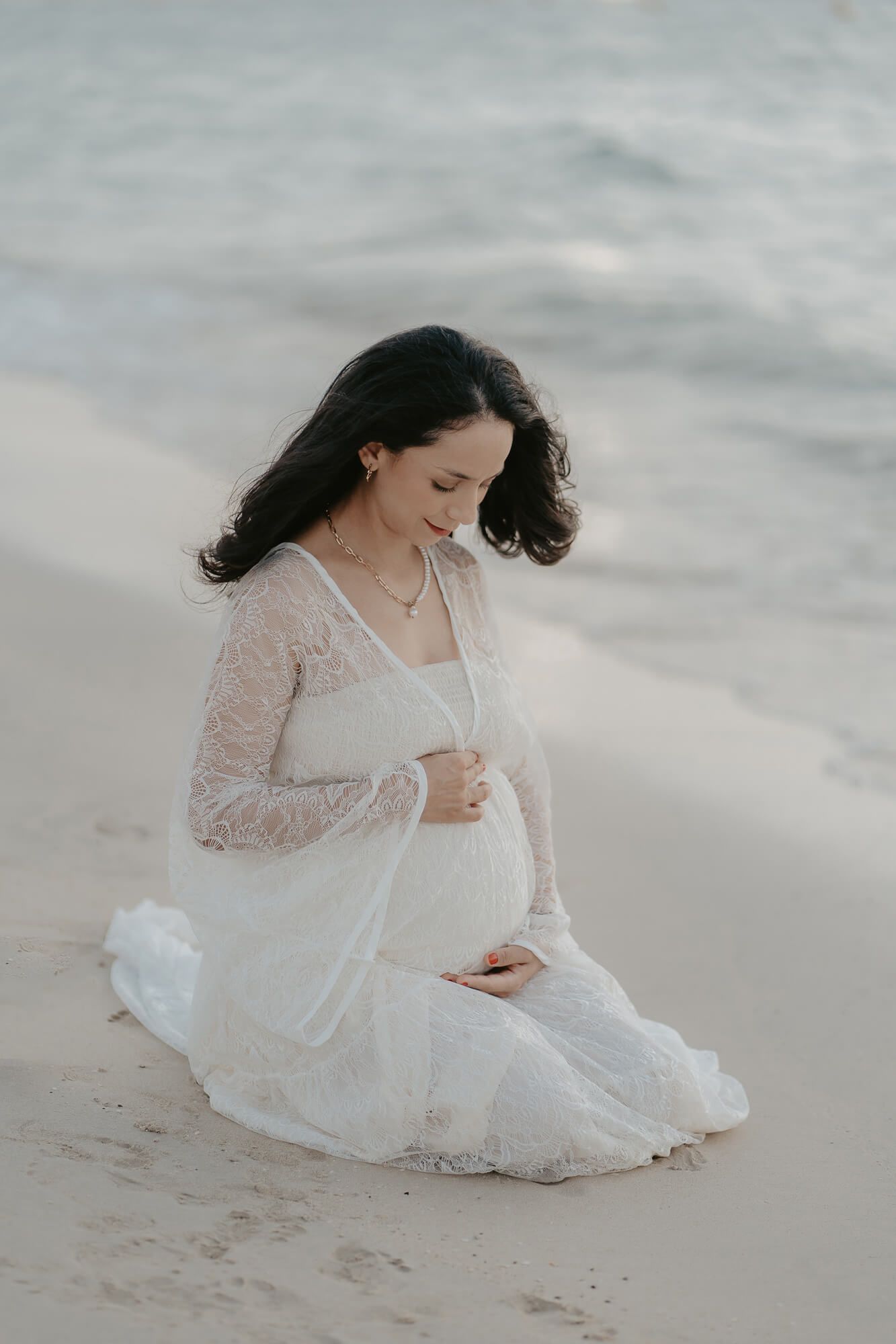 A woman in a flowing white lace dress sits gracefully on a sandy beach in soft, ethereal lighting.
