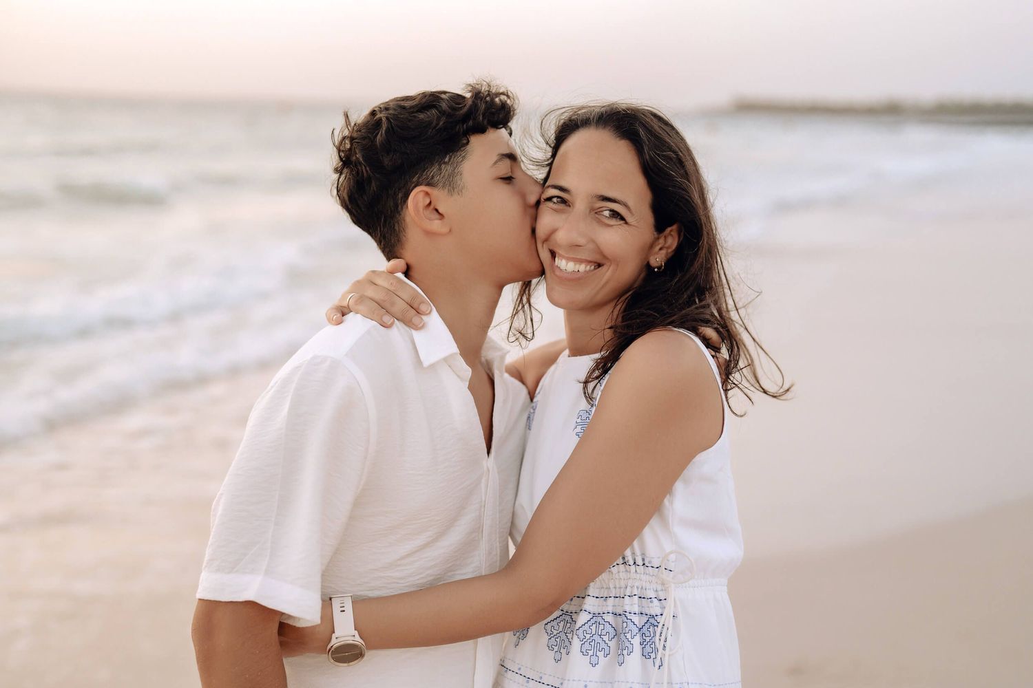 A tender beach photoshoot of two people dressed in white embracing while ocean waves roll in the background.