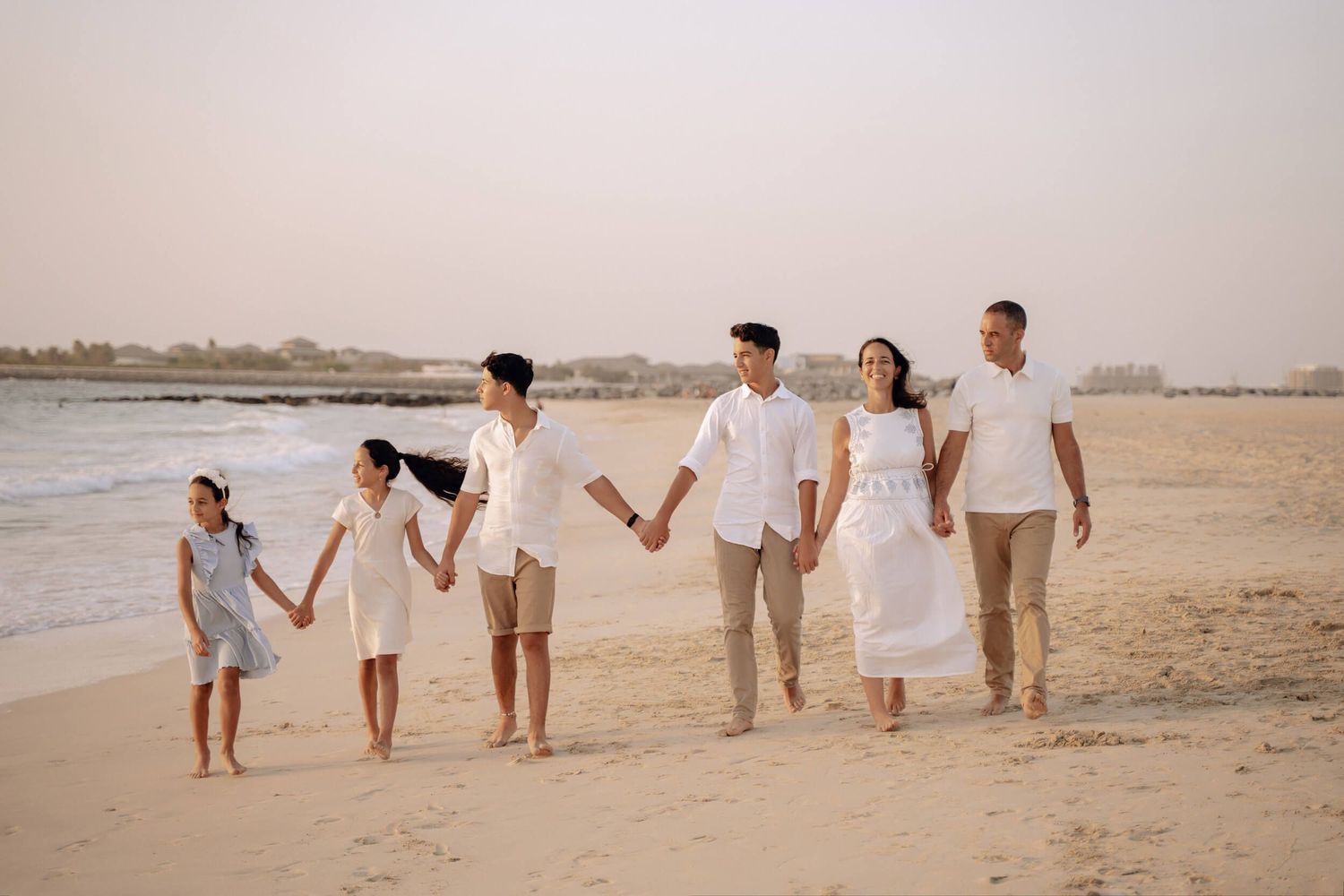 Family walks hand in hand along shoreline wearing coordinated white and neutral outfits.