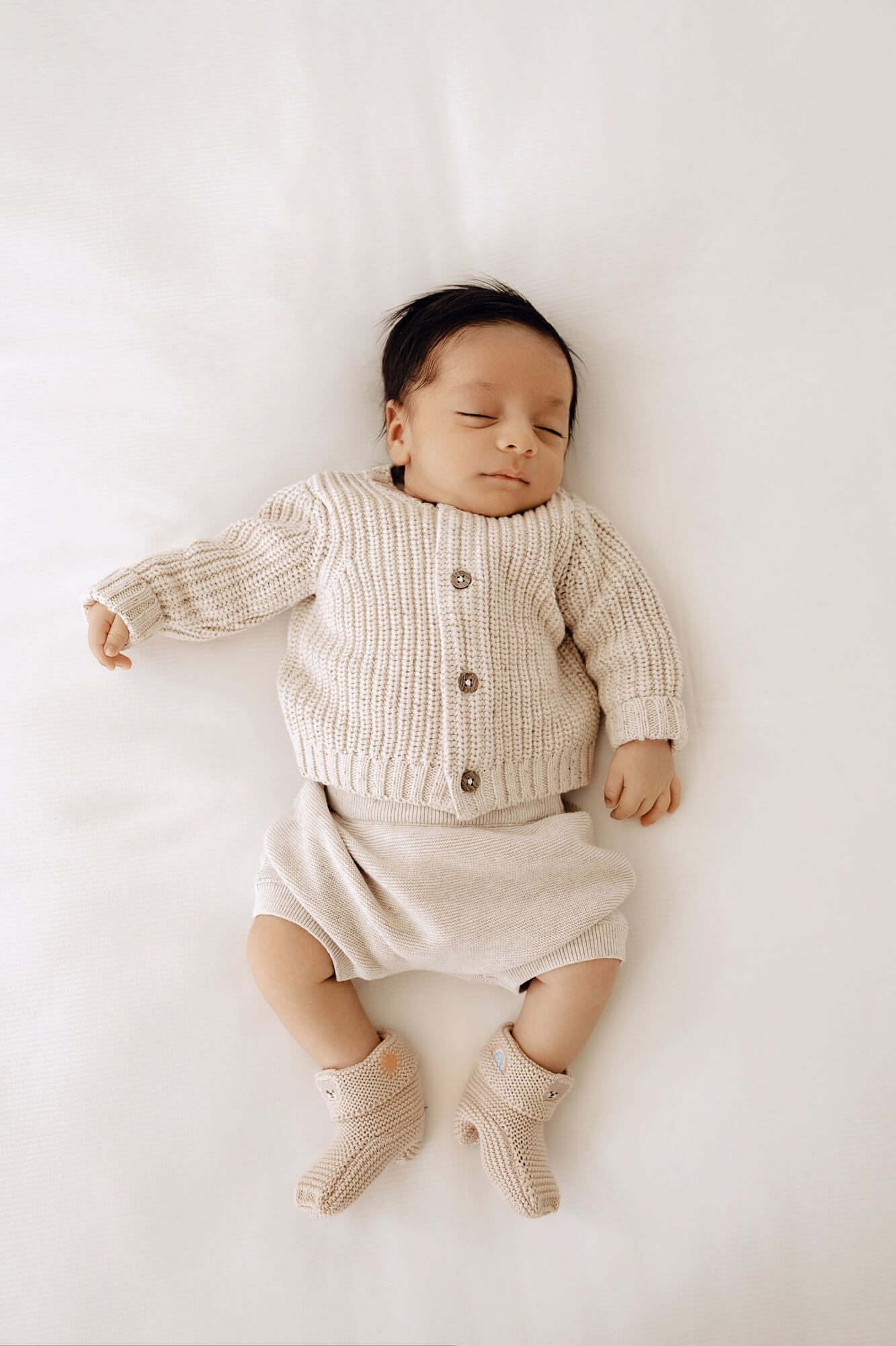 Sleeping baby wearing a cream knit cardigan and shorts lies peacefully on white bedding.