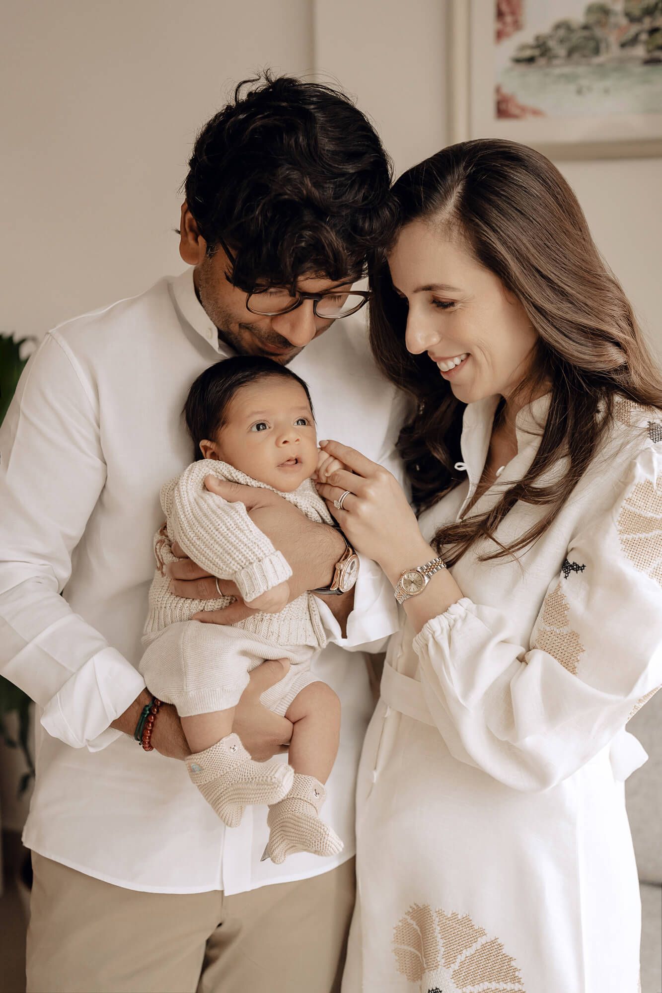 Parents in white clothing share an intimate moment with their baby while standing by a decorative wall.
