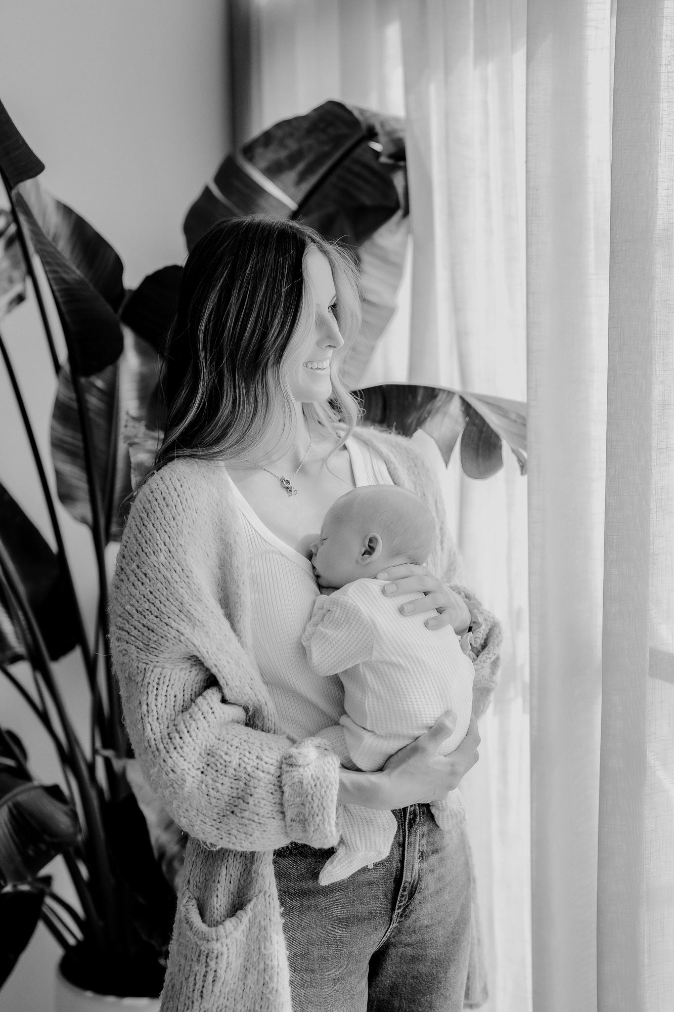 A tender moment captured in black and white as a parent embraces and nurses a baby near a curtained window.