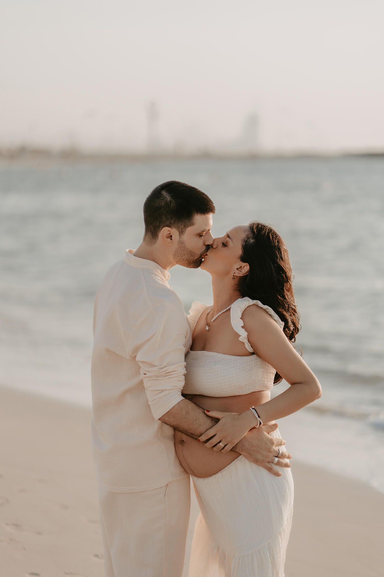 Romantic couple shares an intimate moment embracing on a beach at sunset with city skyline in background.
