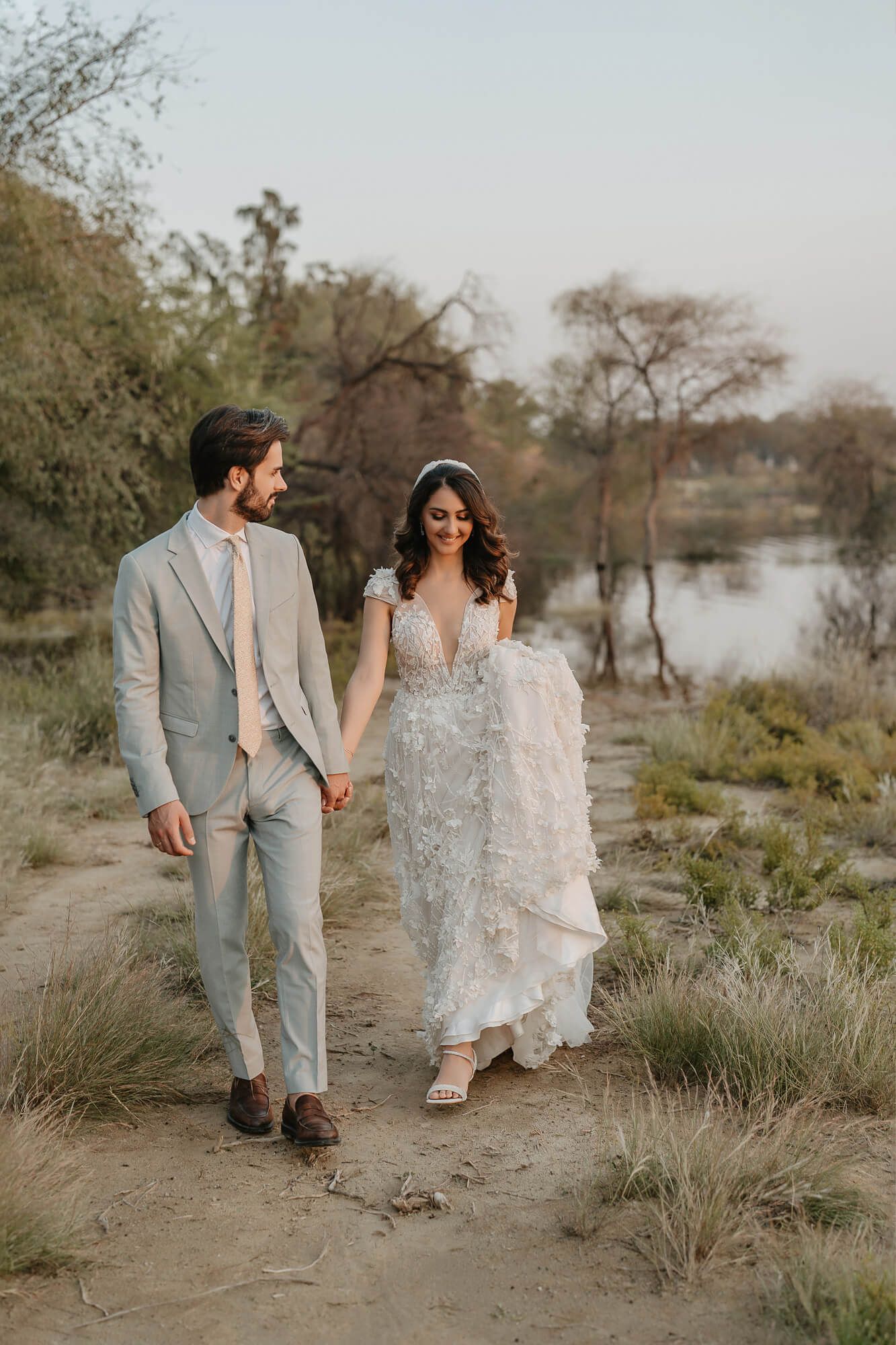 A couple in wedding attire walks through a desert landscape with sparse vegetation at sunset creating a romantic ambiance.