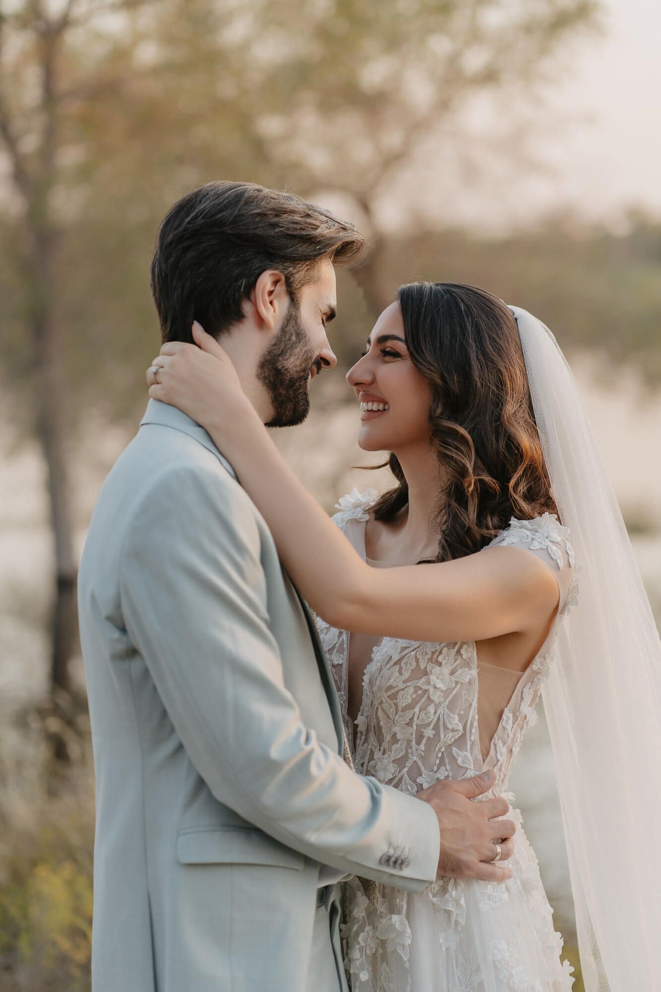 A romantic wedding moment with bride in lace gown and groom in light gray suit embracing outdoors at sunset.