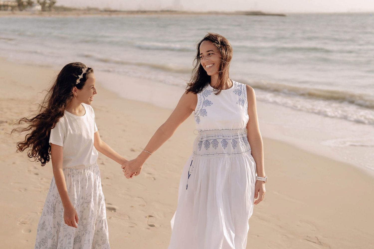 Two children in white dresses holding hands while walking along a beach at sunset.