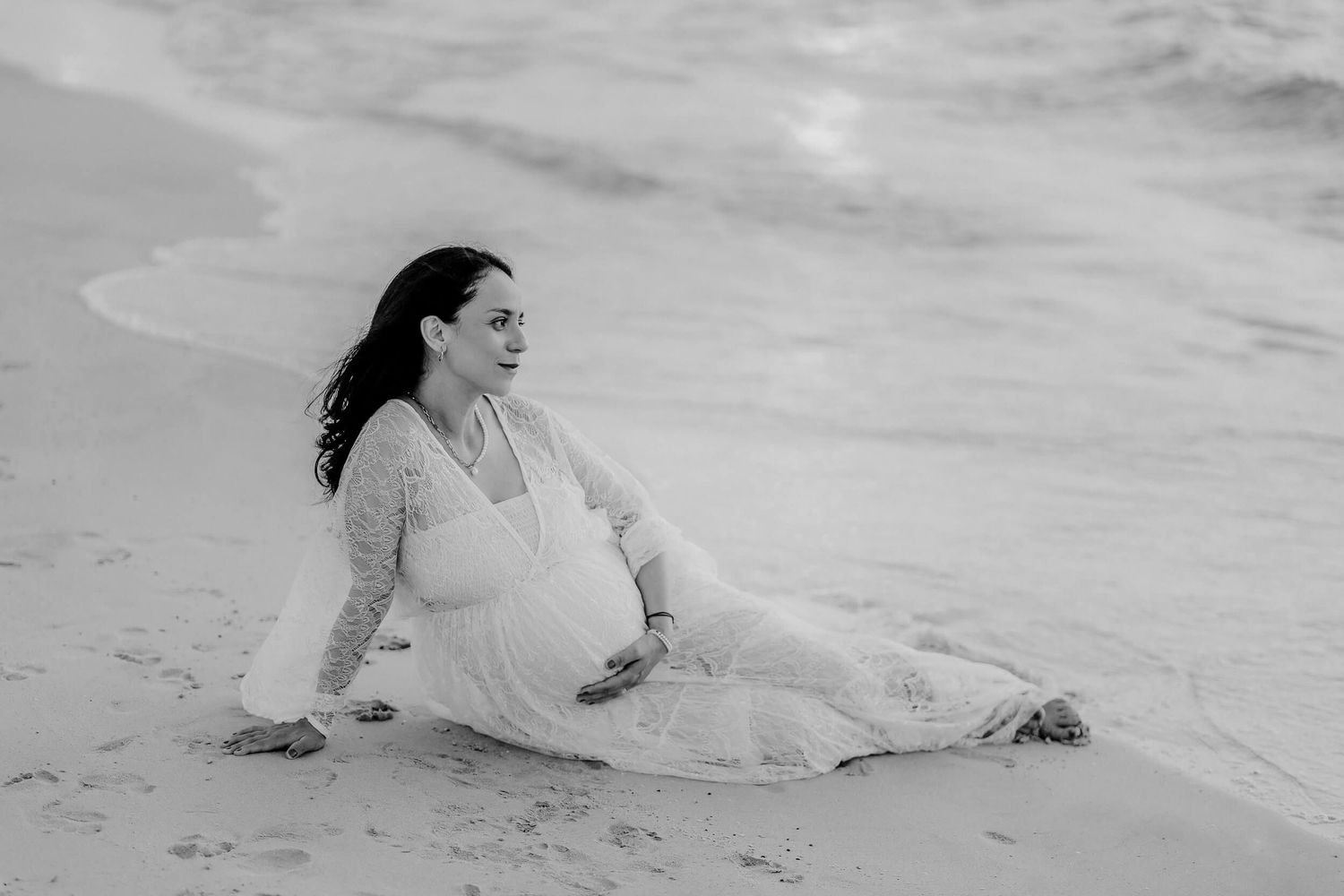 A pregnant woman in a flowing white dress sits peacefully at the edge of ocean waves in a black and white photograph.