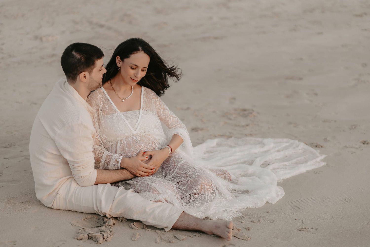 A couple in white outfits sits together on a sandy beach, with the bride's lace dress flowing beside them.
