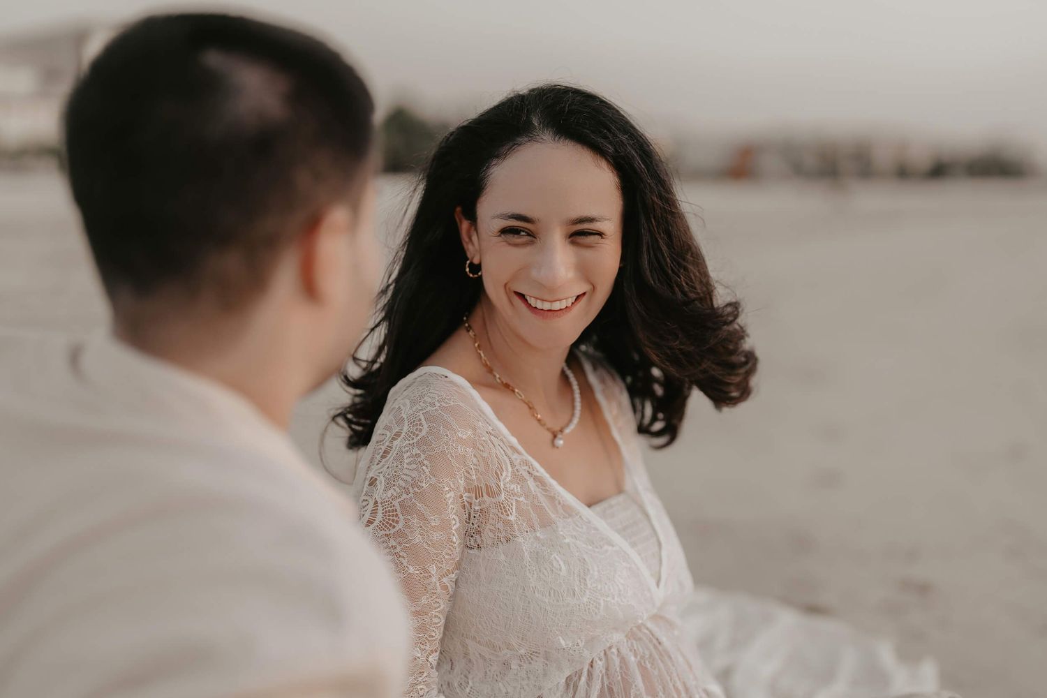 A woman smiling in elegant lace wedding dress poses along beachfront during sunset photoshoot.