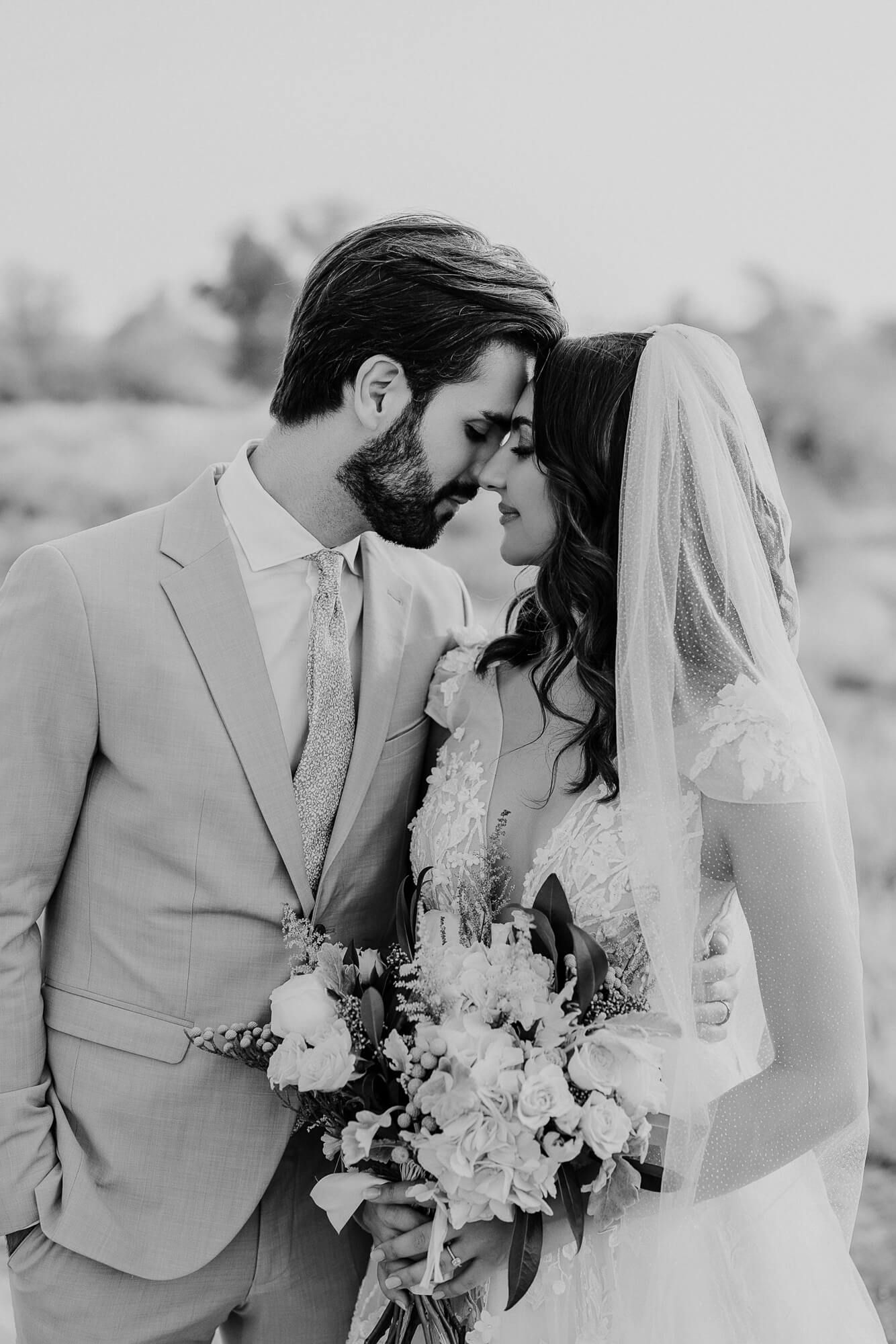 A romantic black and white wedding photograph of a couple sharing an intimate moment while holding a floral bouquet.