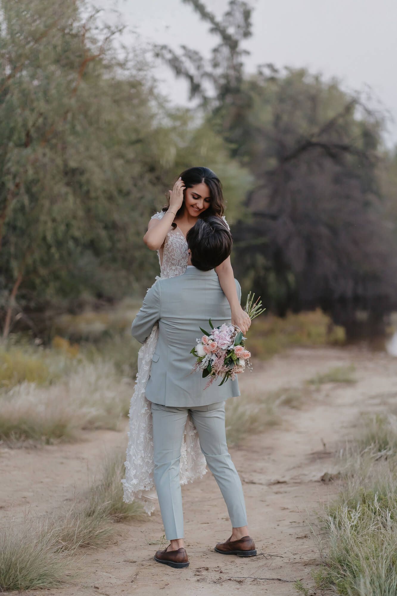 A couple embraces on a dirt path, with the bride holding a pink and white bouquet in a natural outdoor setting.