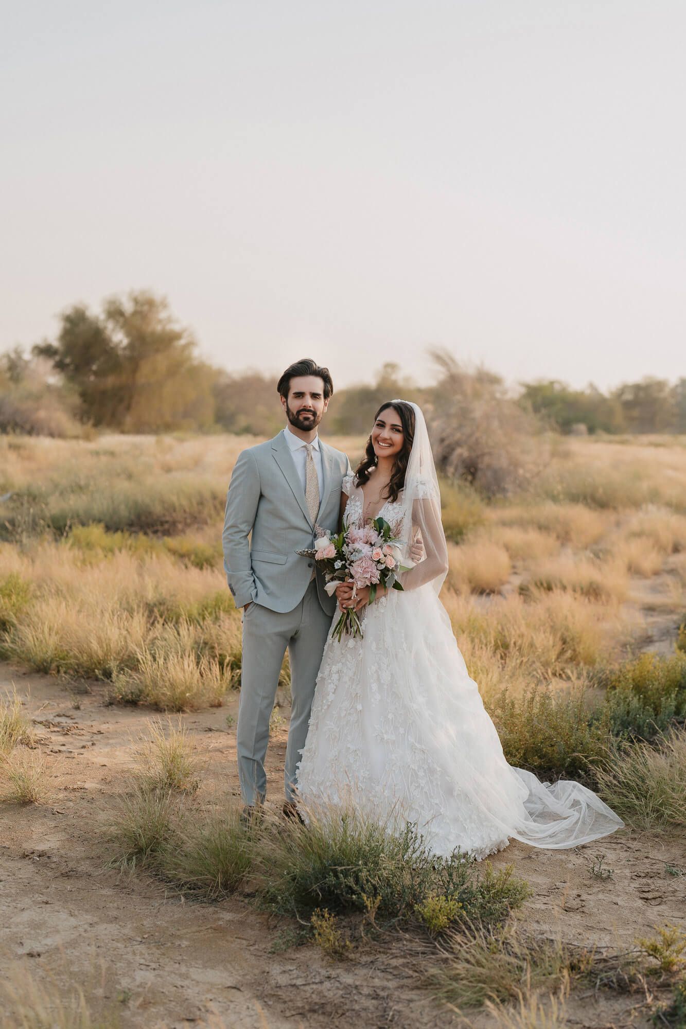 A bride in white lace dress and groom in light gray suit pose together in a desert landscape at sunset.