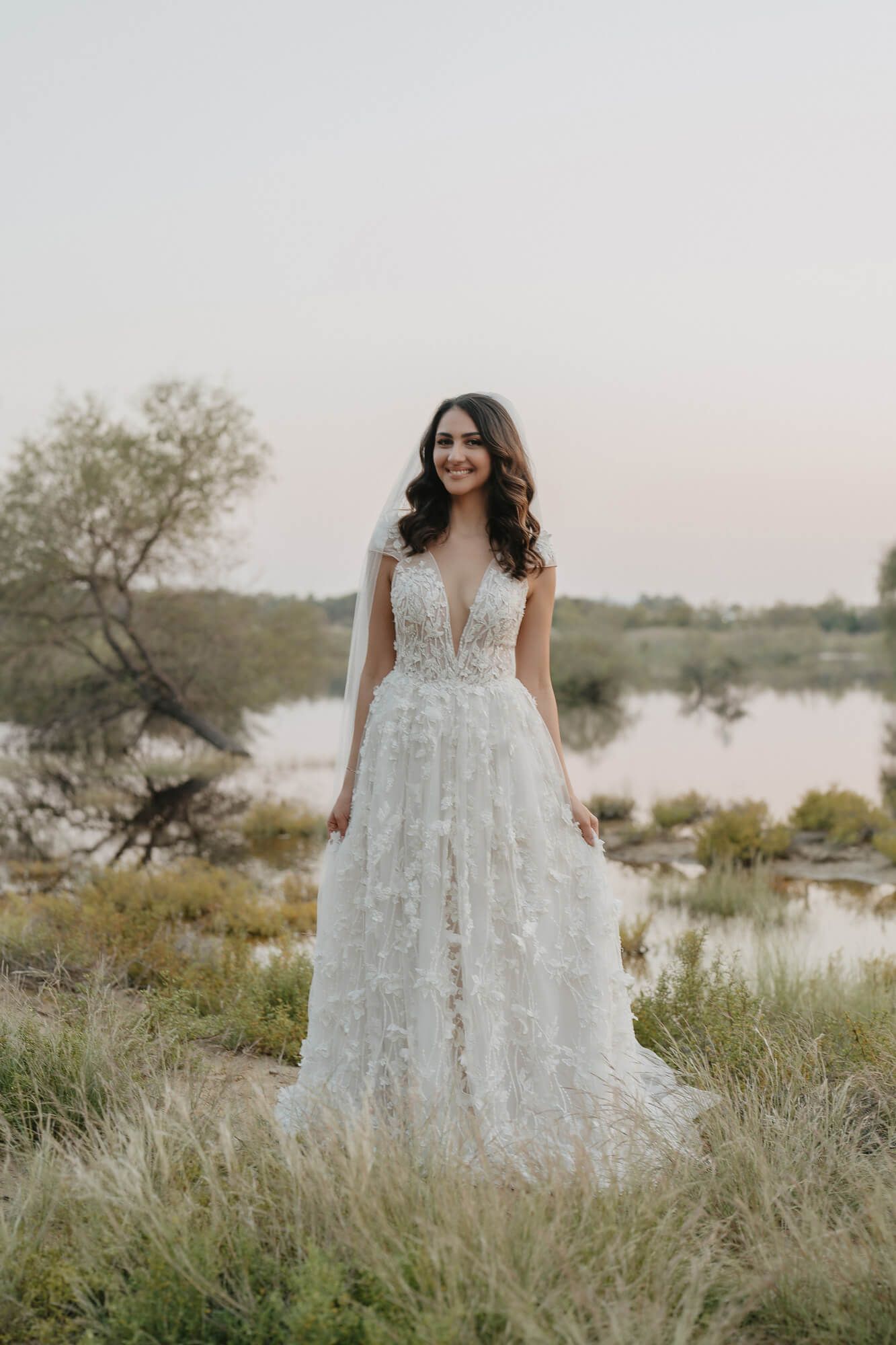 A person in a textured white wedding gown stands gracefully among tall grasses beside a serene lake at sunset.