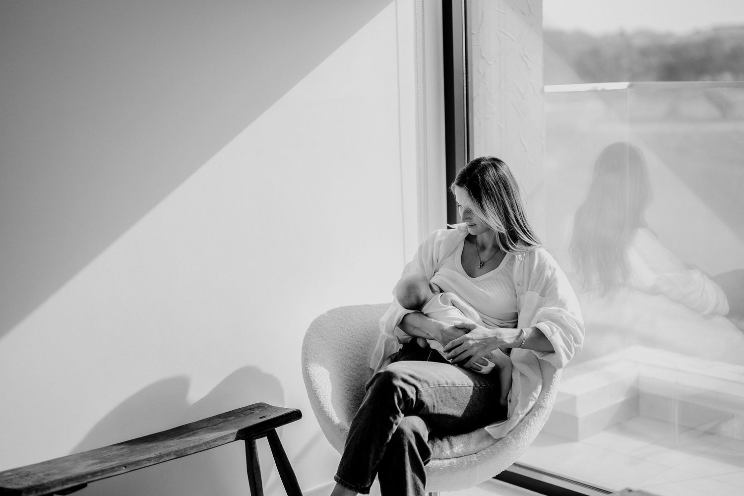 Mother in white sweater cradles baby while sitting by window in black and white photo.