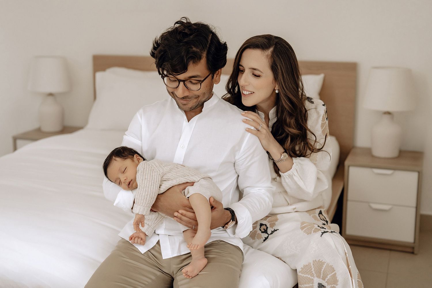 A family shares a tender moment together on a bed with their newborn baby while wearing matching white outfits.