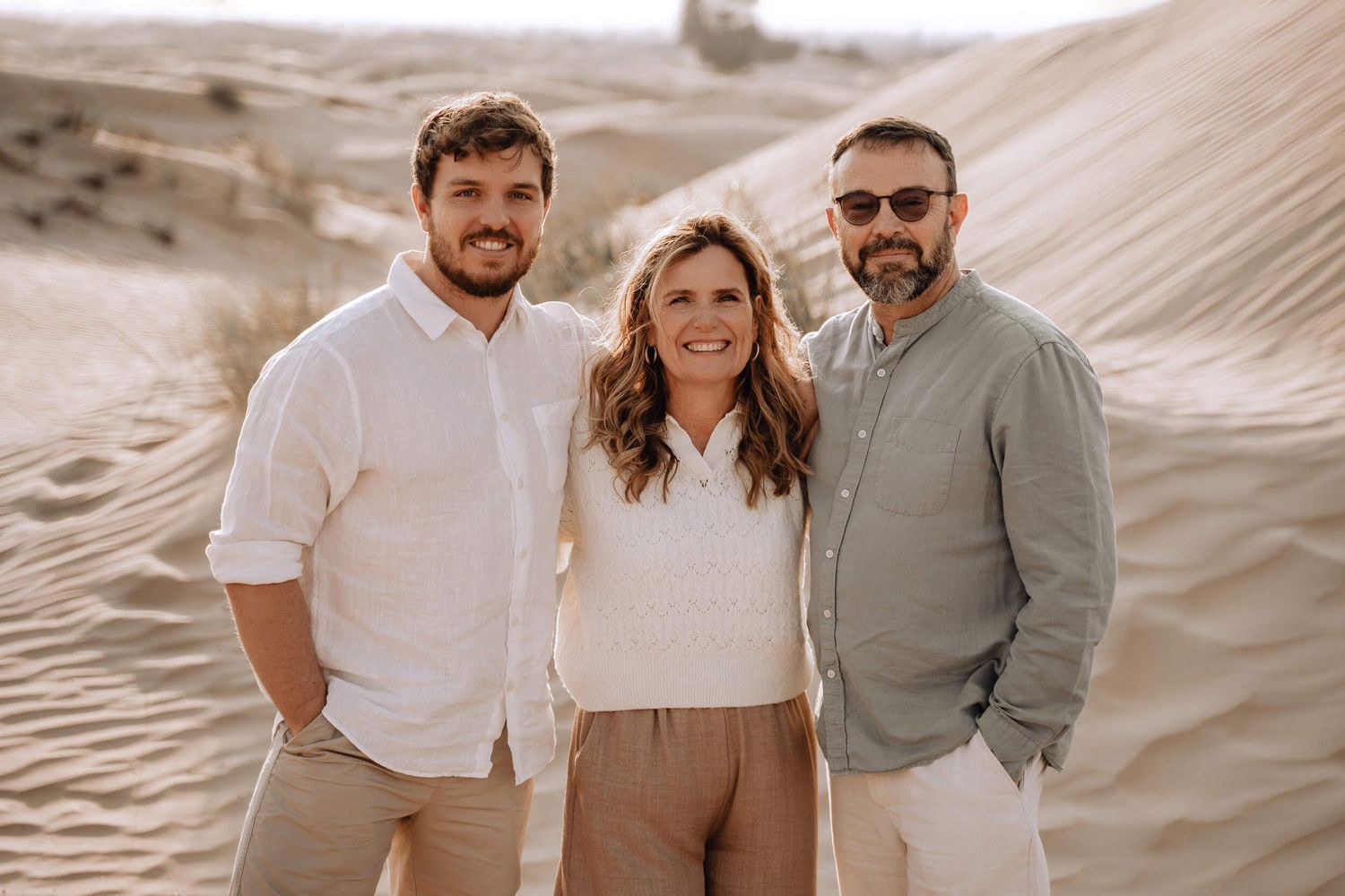 Three people pose together in coordinating outfits against sandy desert dunes backdrop.