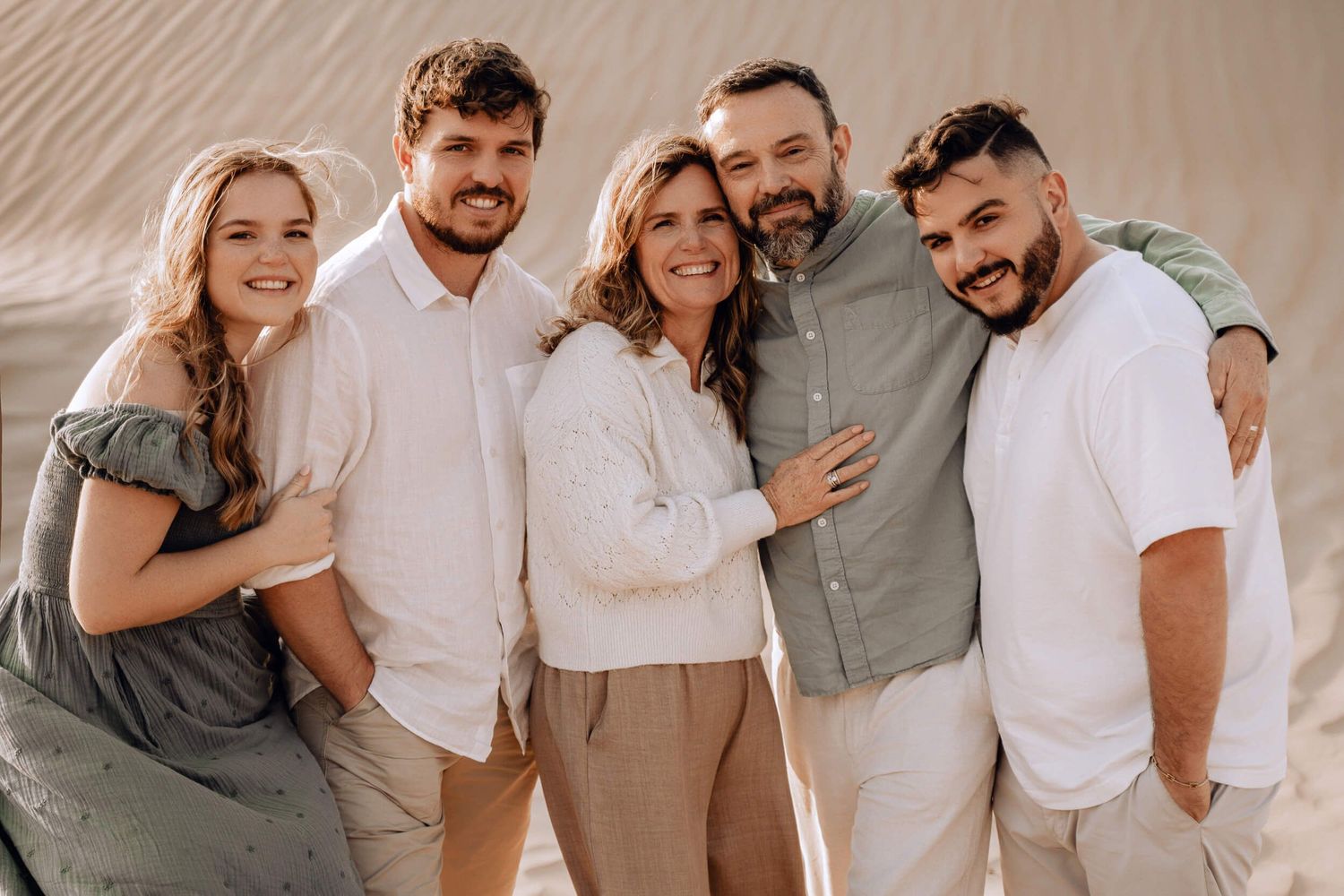 Warm-toned portrait of five people in coordinated outfits embracing on sand dunes.