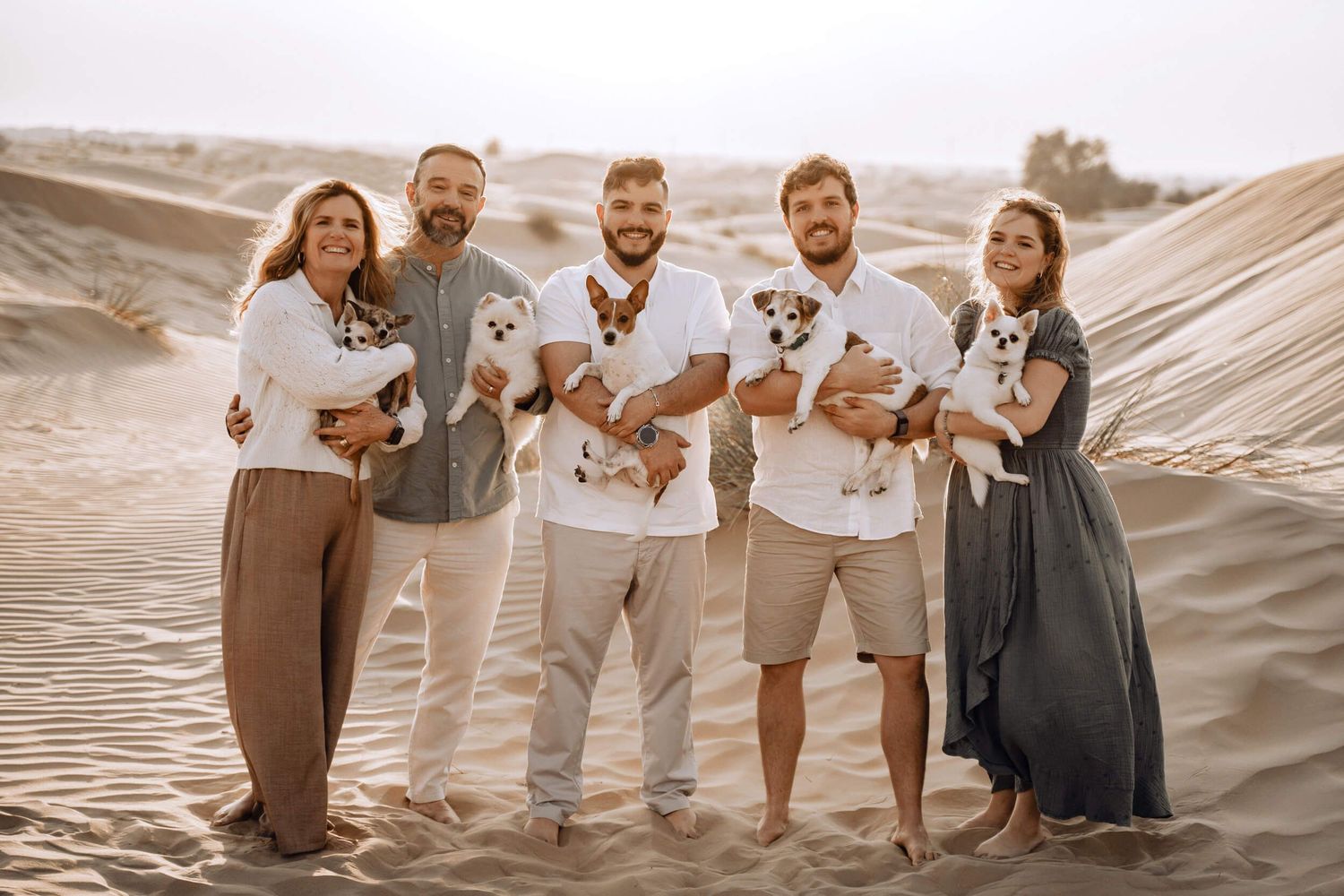 Five people standing together on desert sand holding small puppies in warm-toned photography.