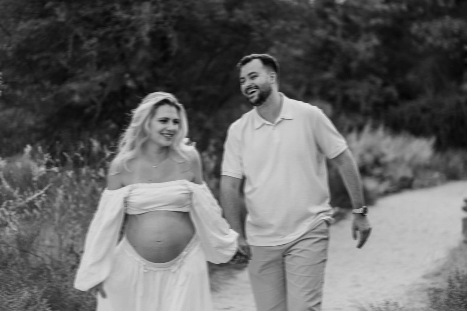 Black and white photo of expecting couple sharing a laugh while walking outdoors.