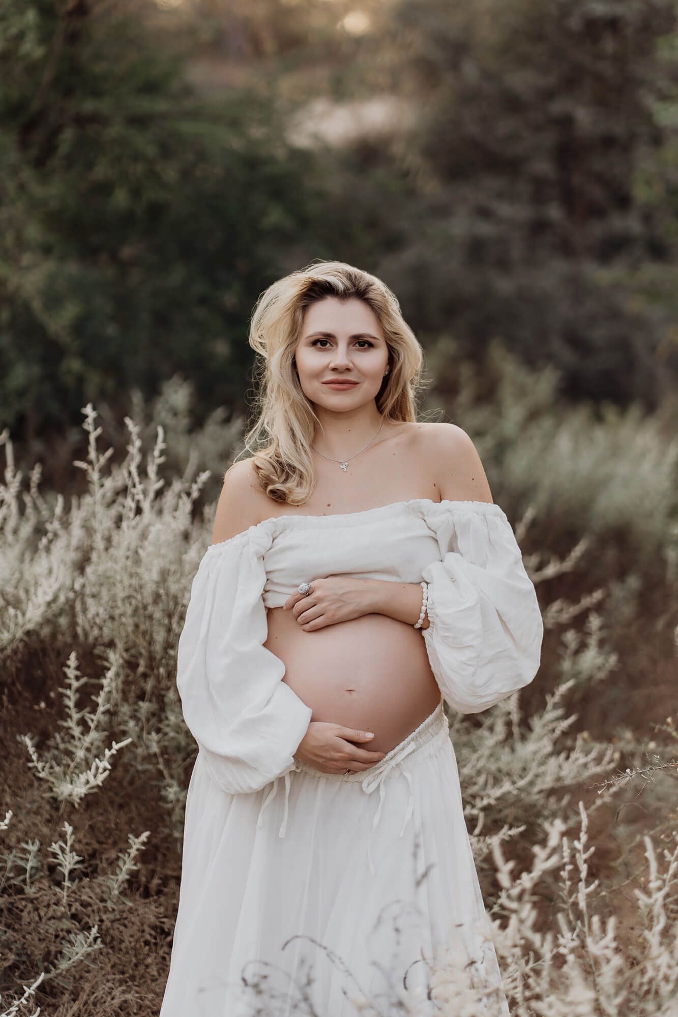 A maternity photoshoot in a field with white flowing dress against a natural outdoor backdrop.