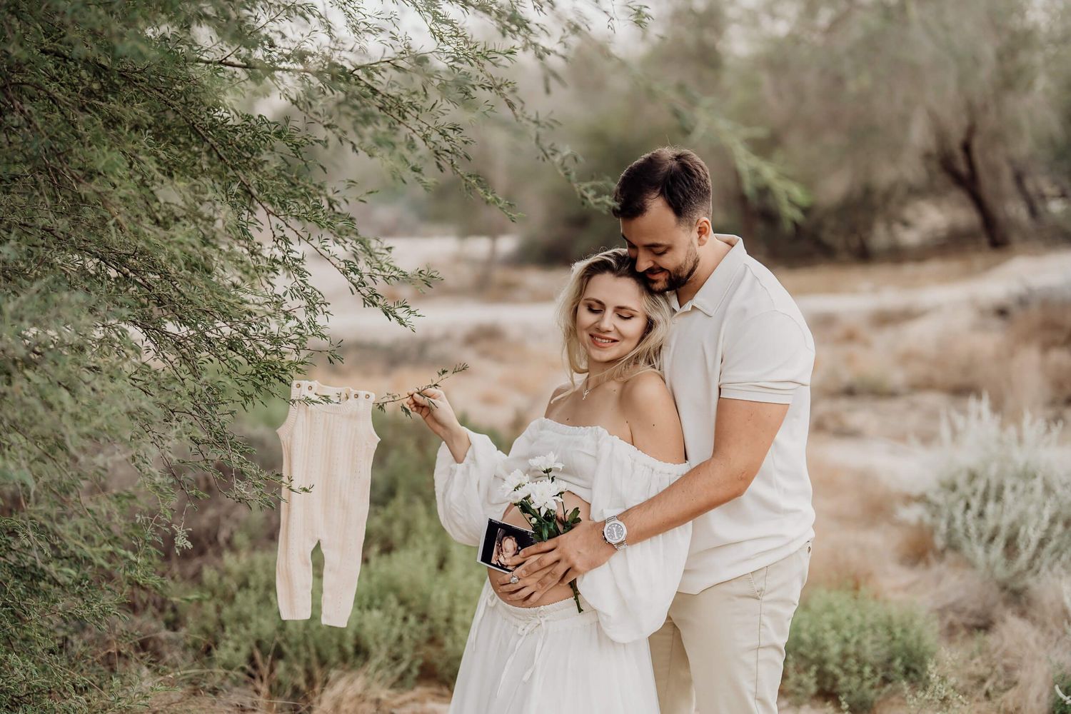 Couple in white outfits share tender moment while holding baby clothes outdoors.