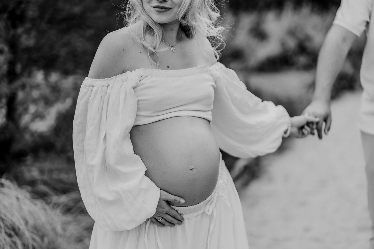 Black and white close-up of pregnant woman in off-shoulder white dress showing her baby bump.