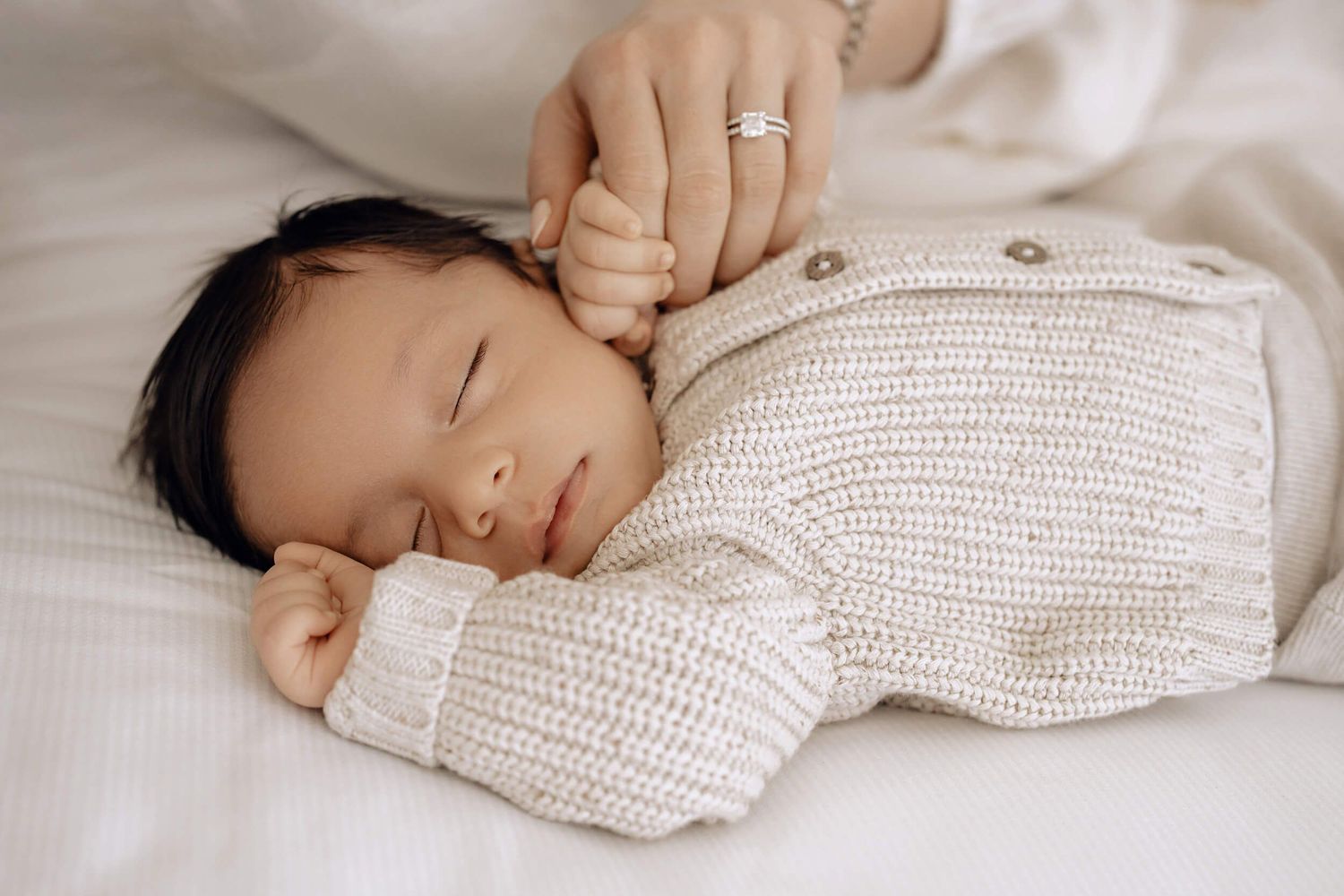 Infant rests comfortably in a cozy cream knit top on white bedding.
