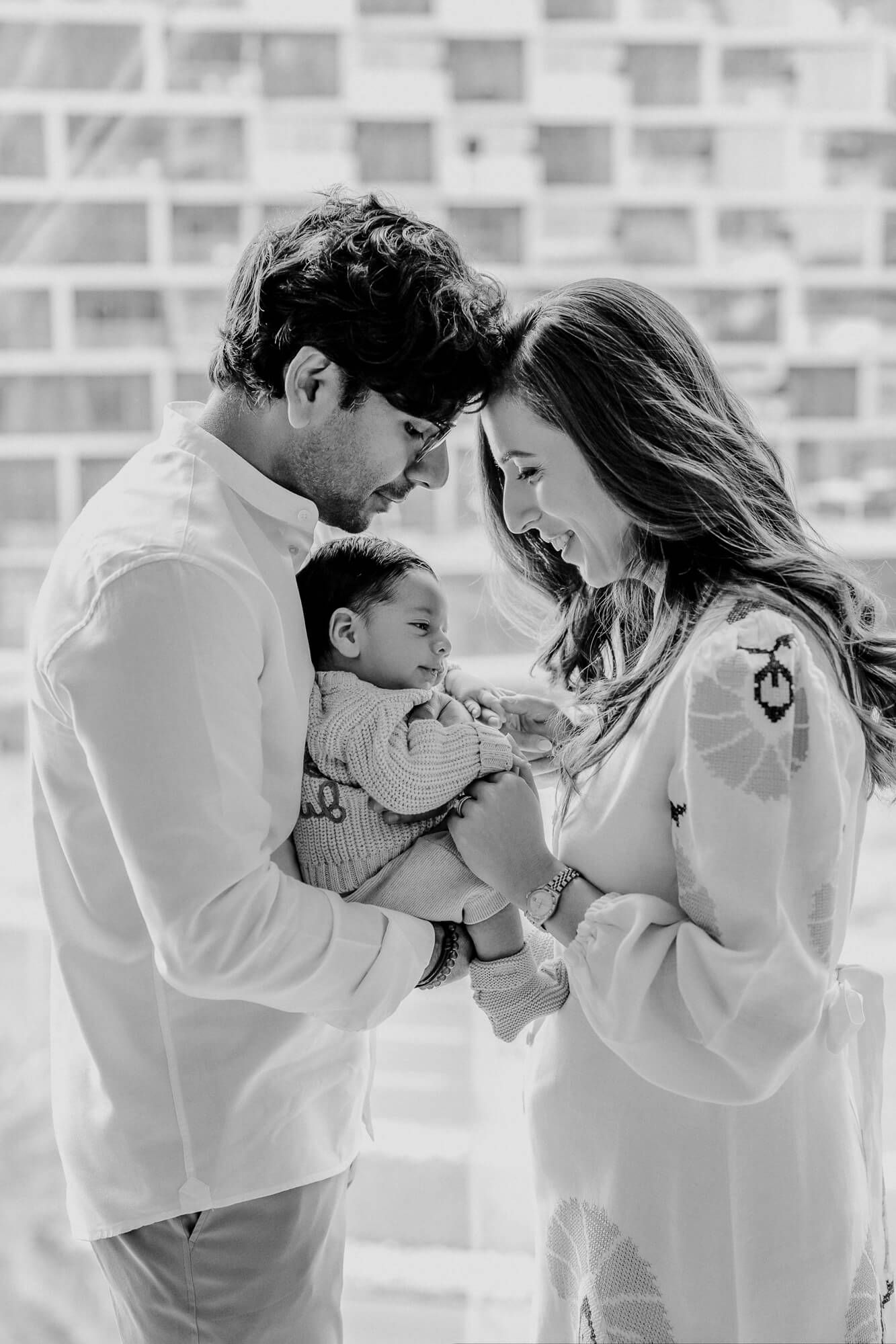 Black and white candid portrait of parents sharing joyful moment with baby against geometric wall.