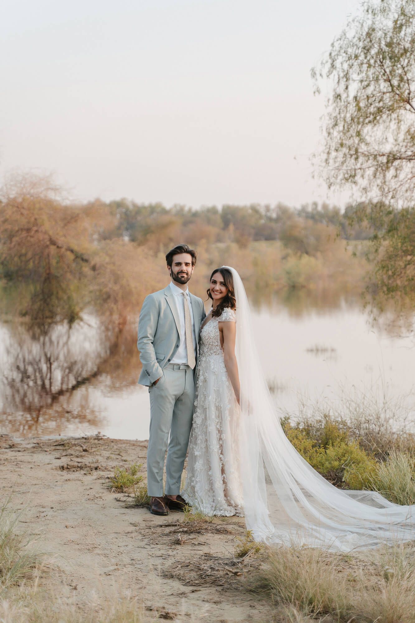 A romantic wedding portrait taken at sunset by a lake with a couple in light blue suit and flowing lace gown.
