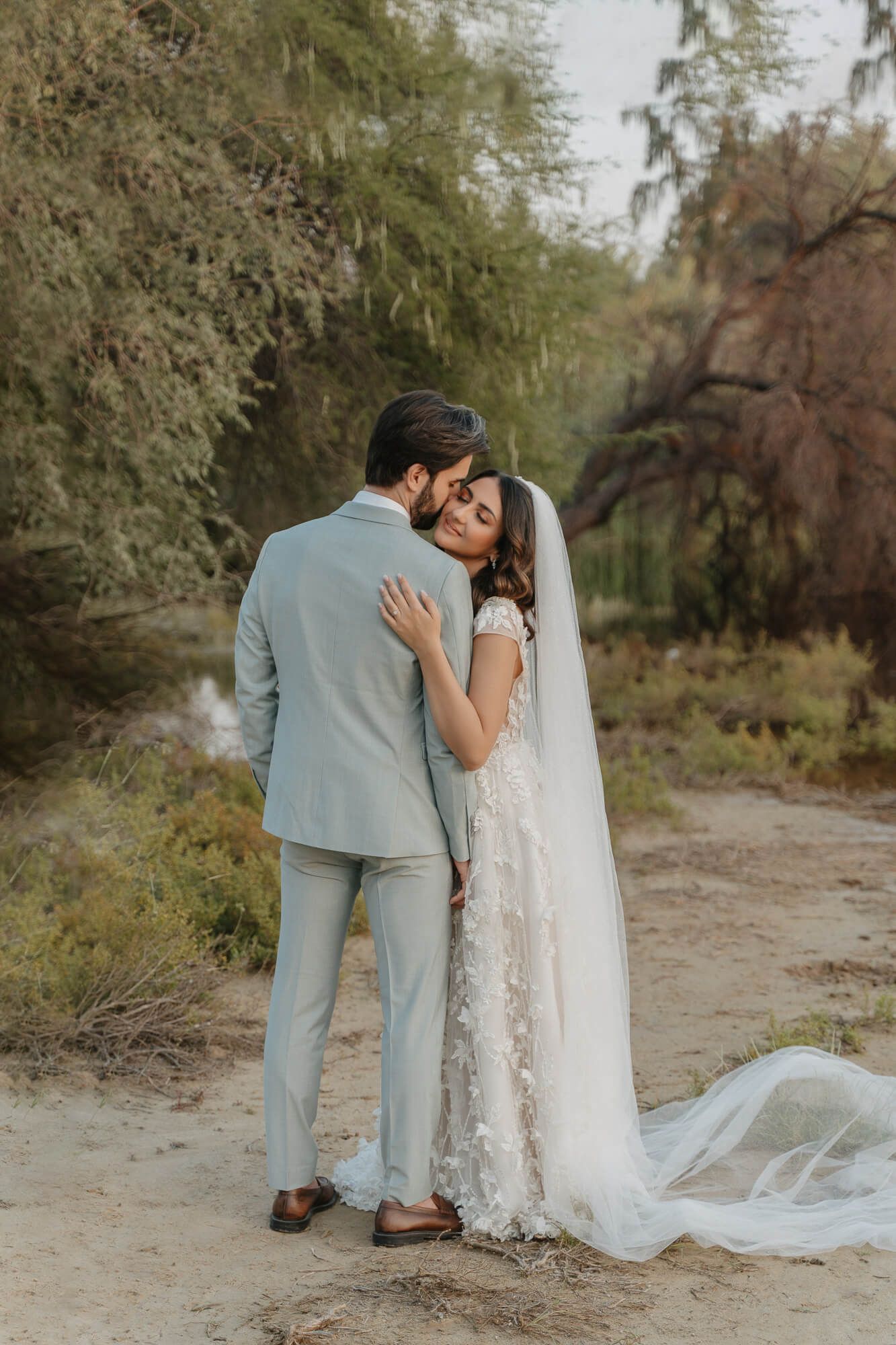 A couple embraces outdoors in wedding attire, with the bride wearing a flowing lace gown and long veil on a nature path.