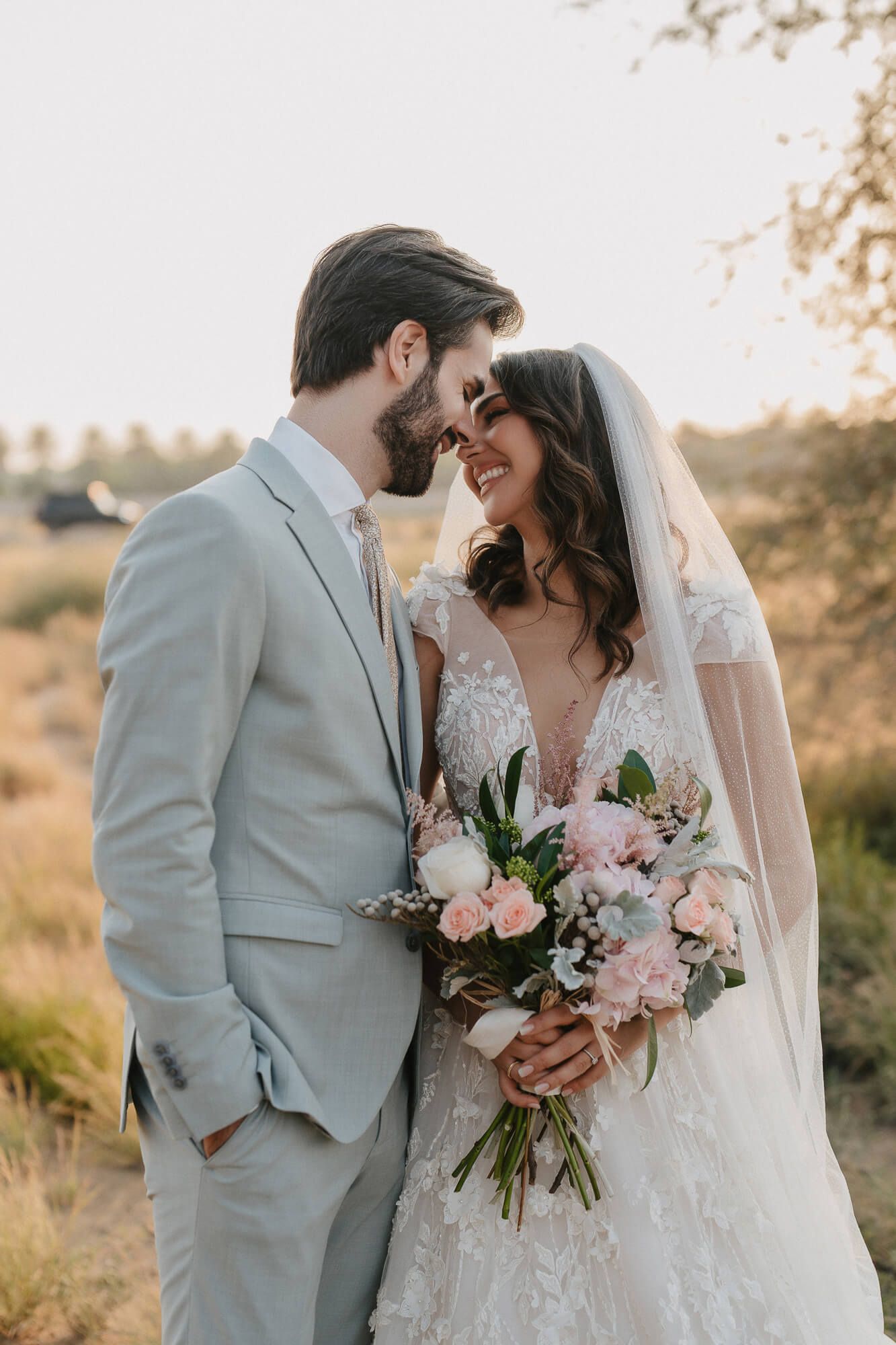 A romantic wedding couple embraces in the golden sunlight holding a pink and green floral bouquet.
