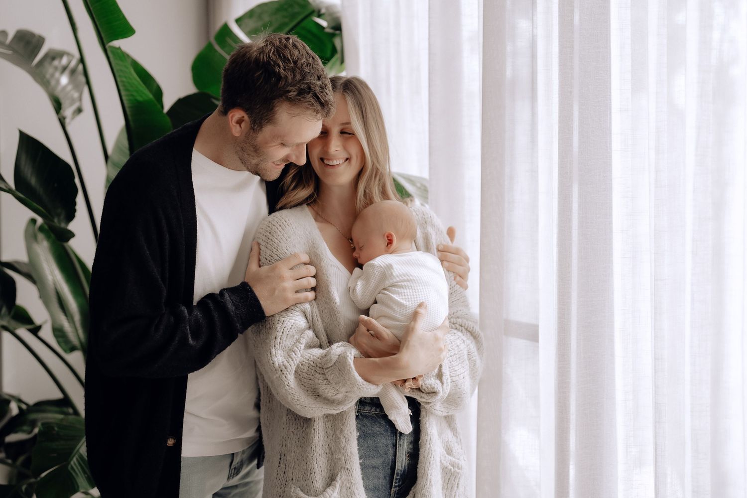 A couple sharing an intimate moment with their baby by a window with white curtains and indoor plants.