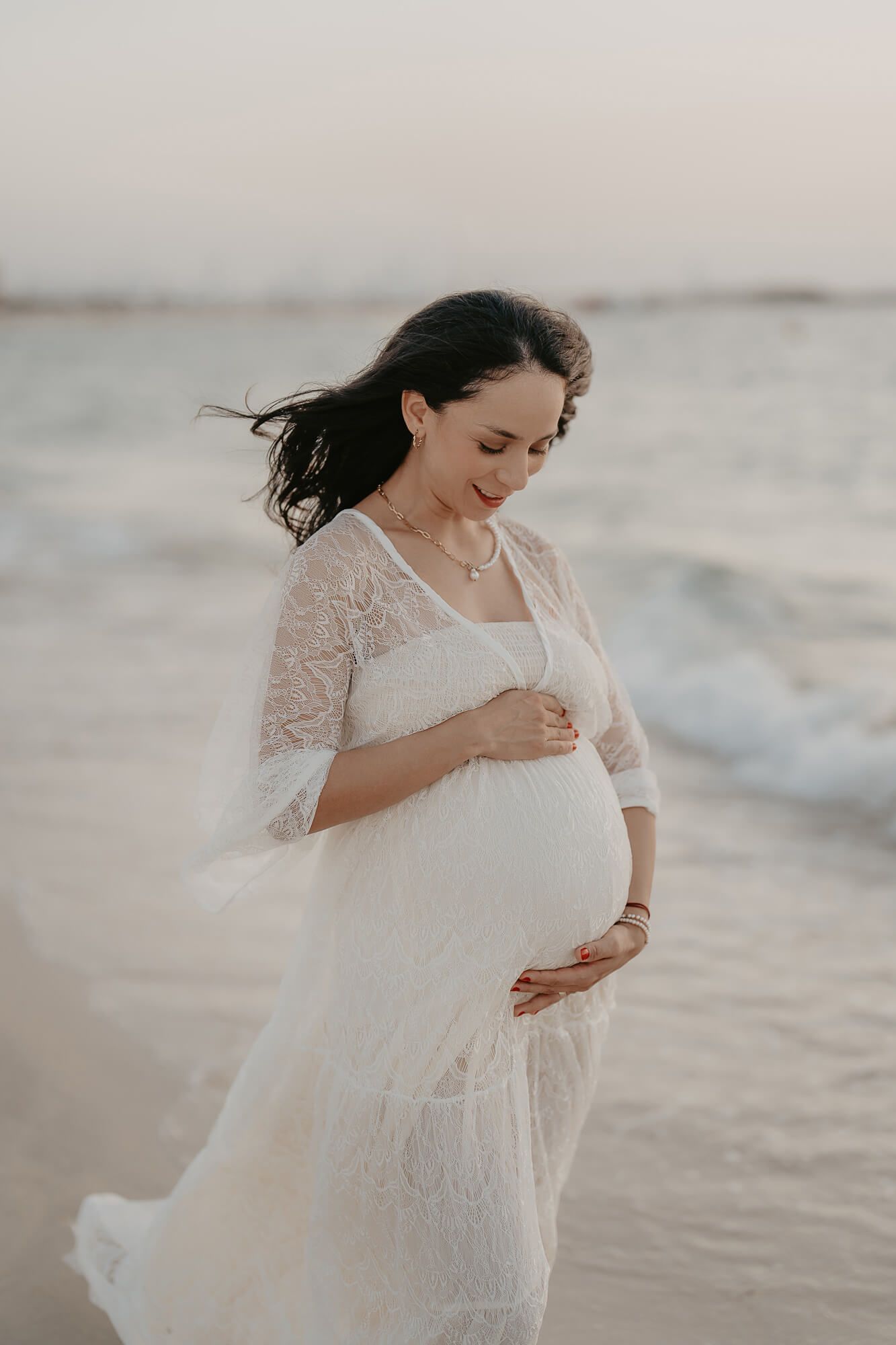 Expecting mother wearing flowing white lace dress walks along beach at sunset with wind in her hair.