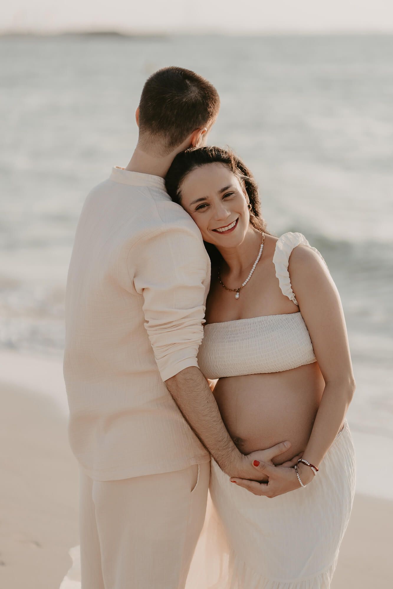 Couple in white outfits sharing an intimate embrace on the beach during a maternity photoshoot.