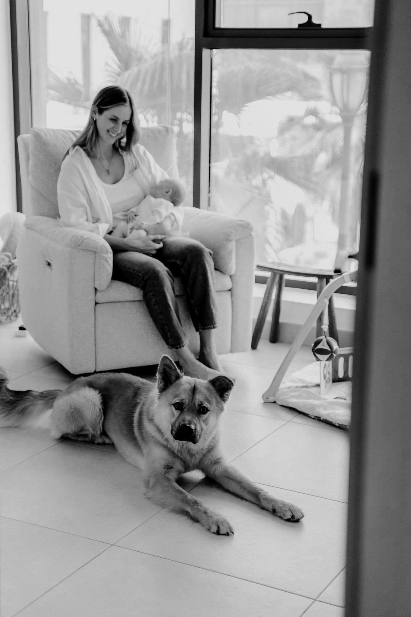 Black and white photo of a quiet moment at home with a large dog resting on tile floor near a comfortable armchair.