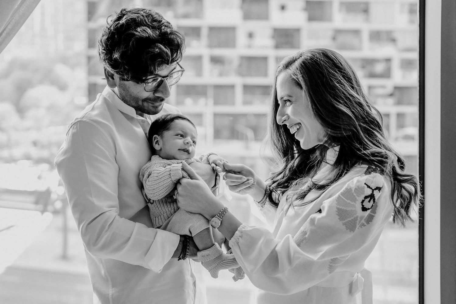 Black and white photo of parents playing with their newborn baby in natural window light.