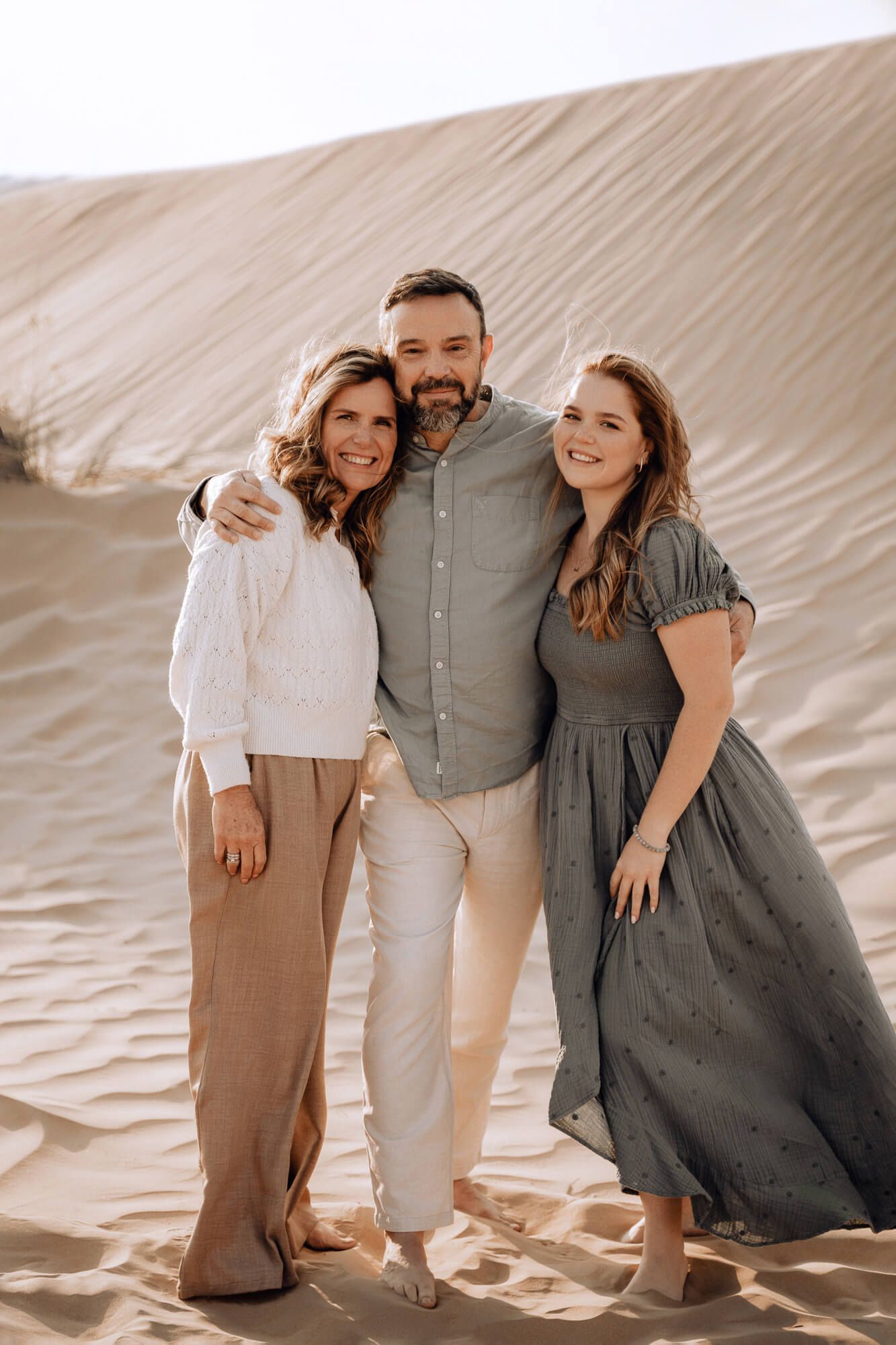A stylish family portrait in coordinated neutral outfits standing together against sand dunes in warm sunlight.