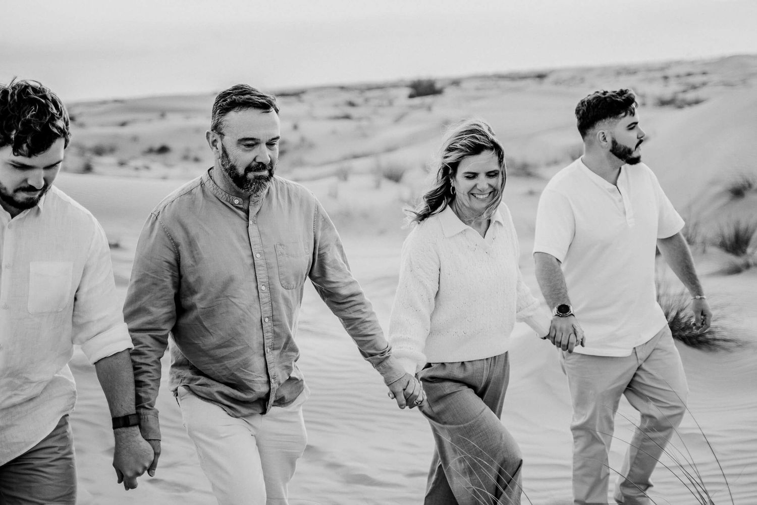 Black and white capture of friends walking together across desert sand dunes in light colored clothing.
