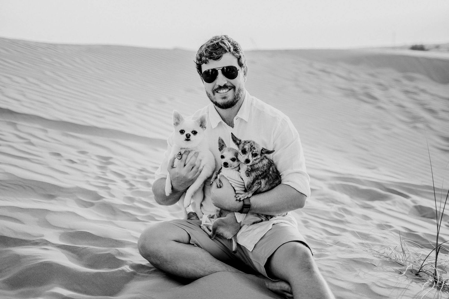 Black and white photo of person with sunglasses on sand dunes holding two small dogs.