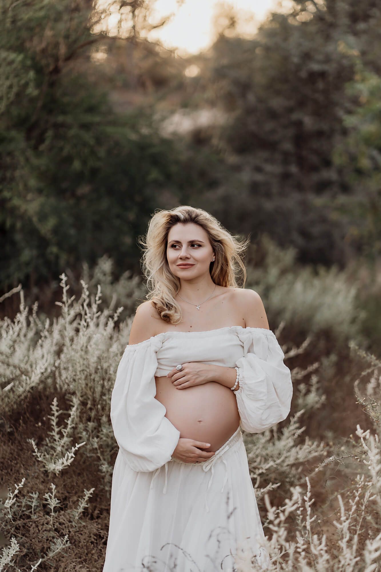 Ethereal maternity portrait in white dress surrounded by wild grasses at sunset.