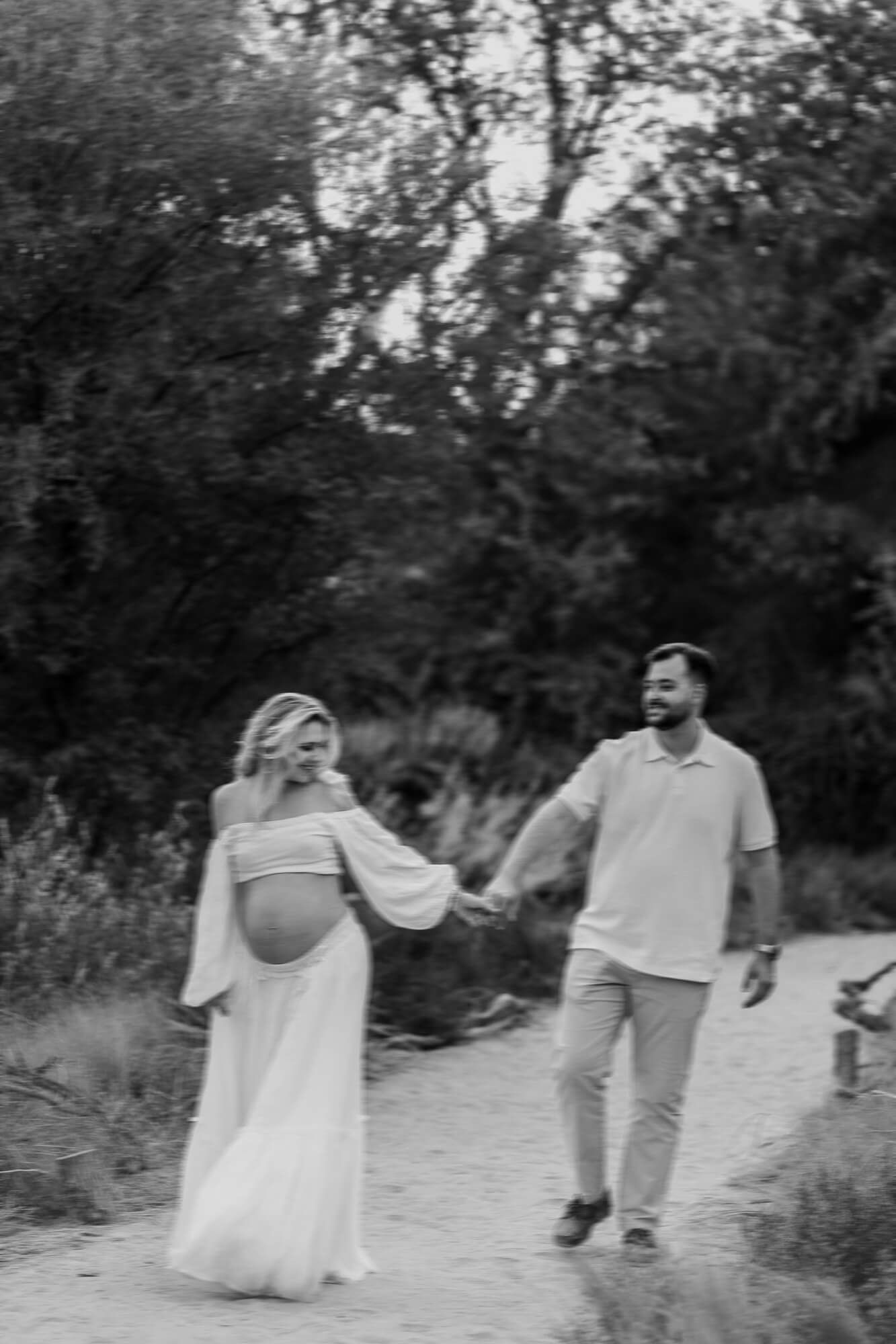Black and white maternity photo of couple walking hand in hand on nature trail.