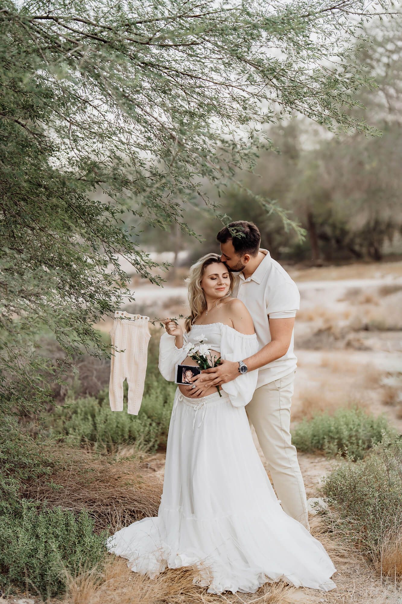 Couple embraces while holding baby clothes and flowers under desert tree branches.