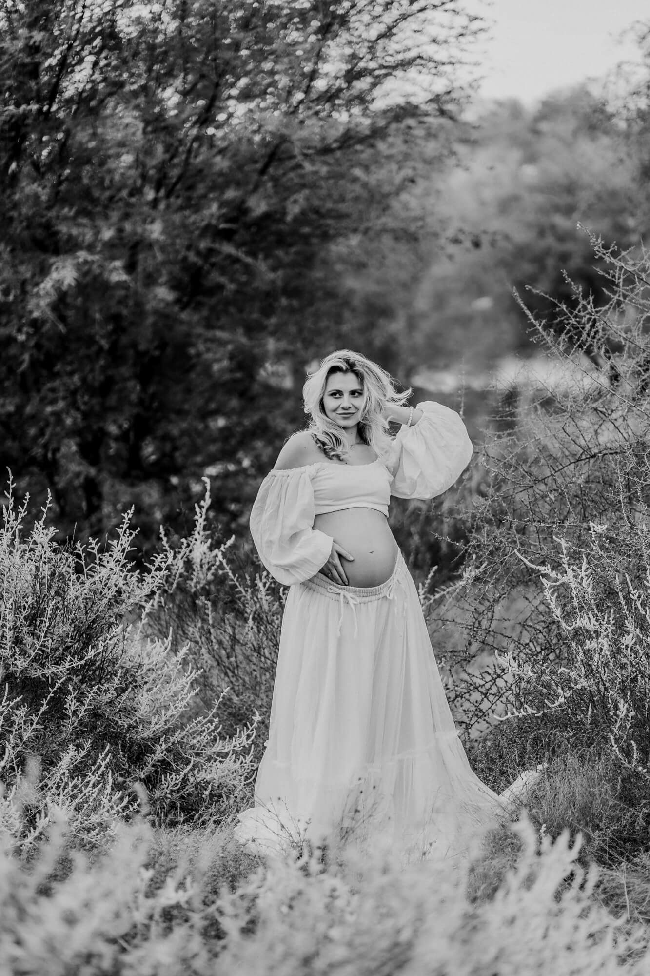 A maternity photoshoot in nature featuring a flowing white dress and soft lighting in a desert landscape.