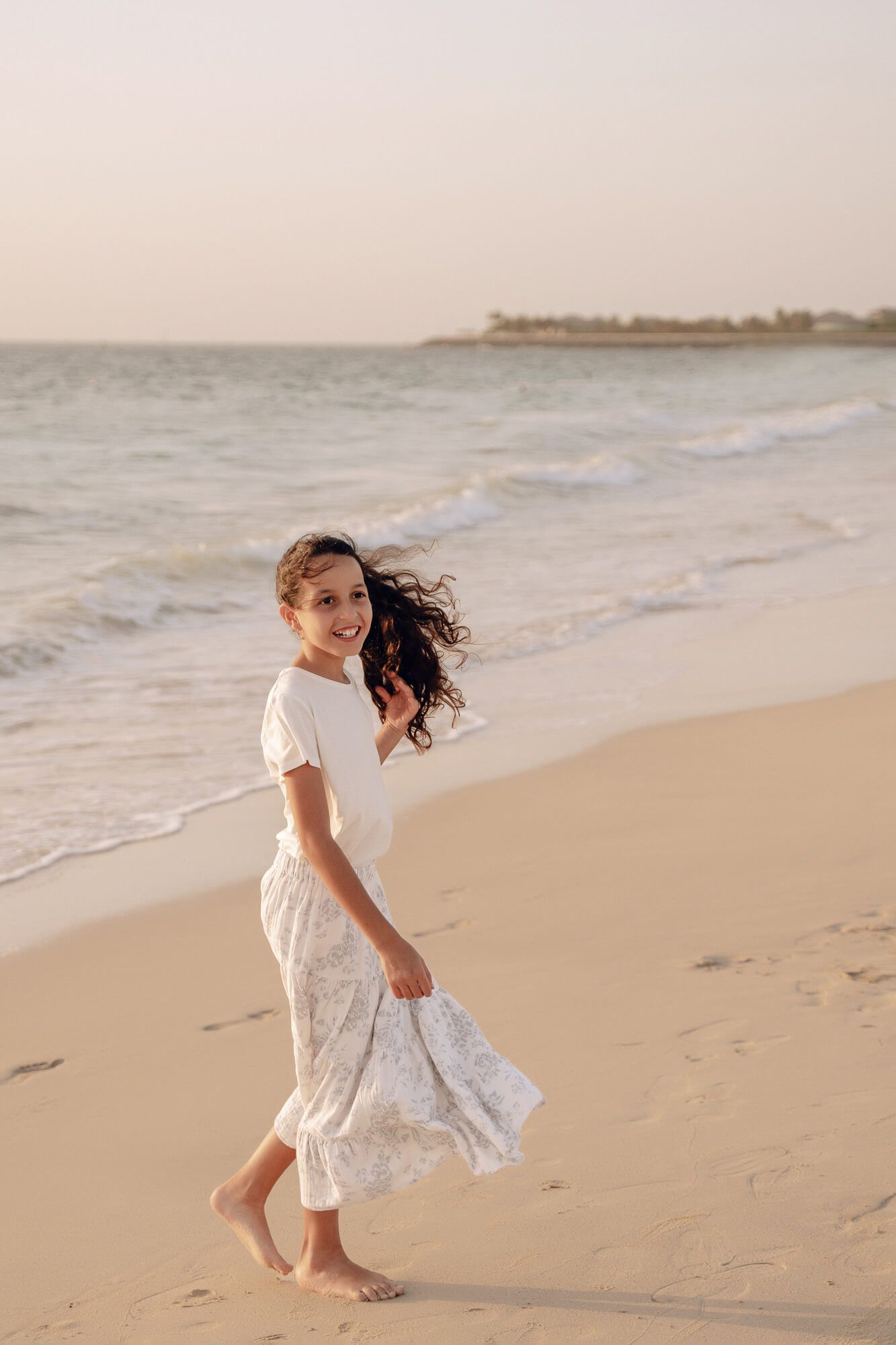 Young girl in a white dress twirls playfully on a sandy beach at sunset.